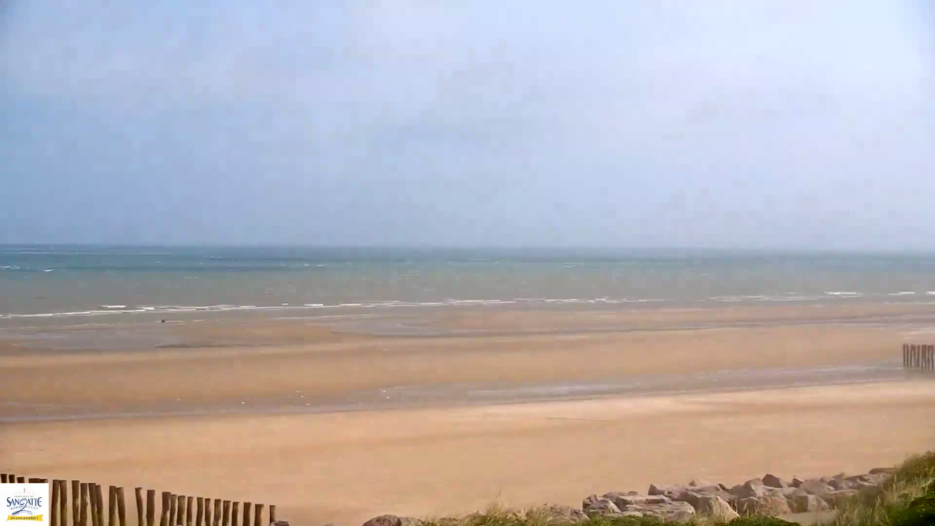 A wide sandy beach stretches to a wavy ocean under a hazy sky.