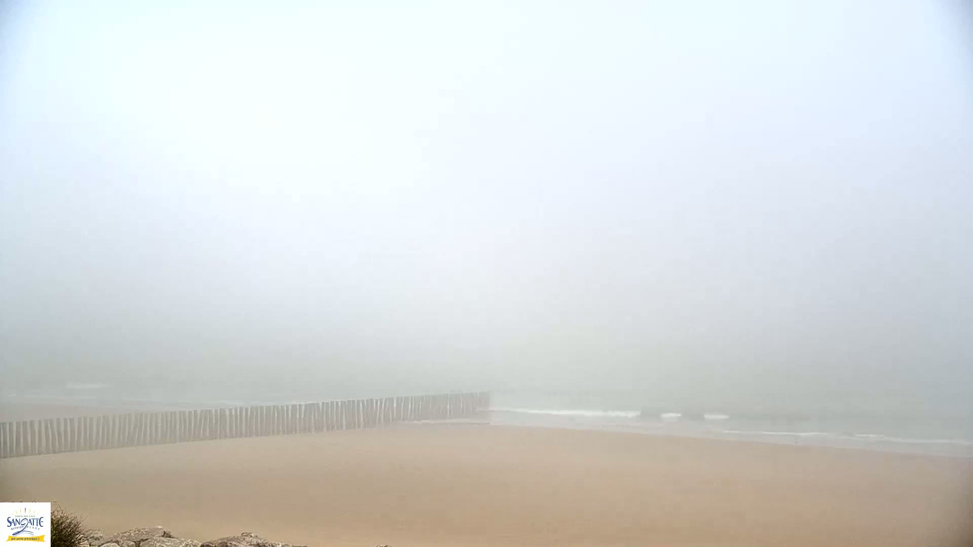 A foggy beach scene shows a sandy shore with a wooden barrier separating the beach from the ocean.