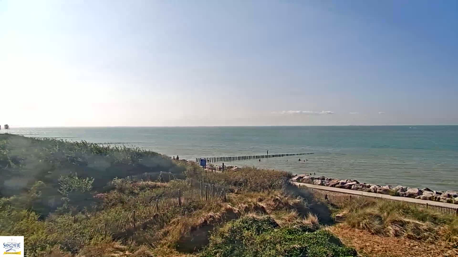 A foggy beach scene shows a sandy shore with a wooden barrier separating the beach from the ocean.