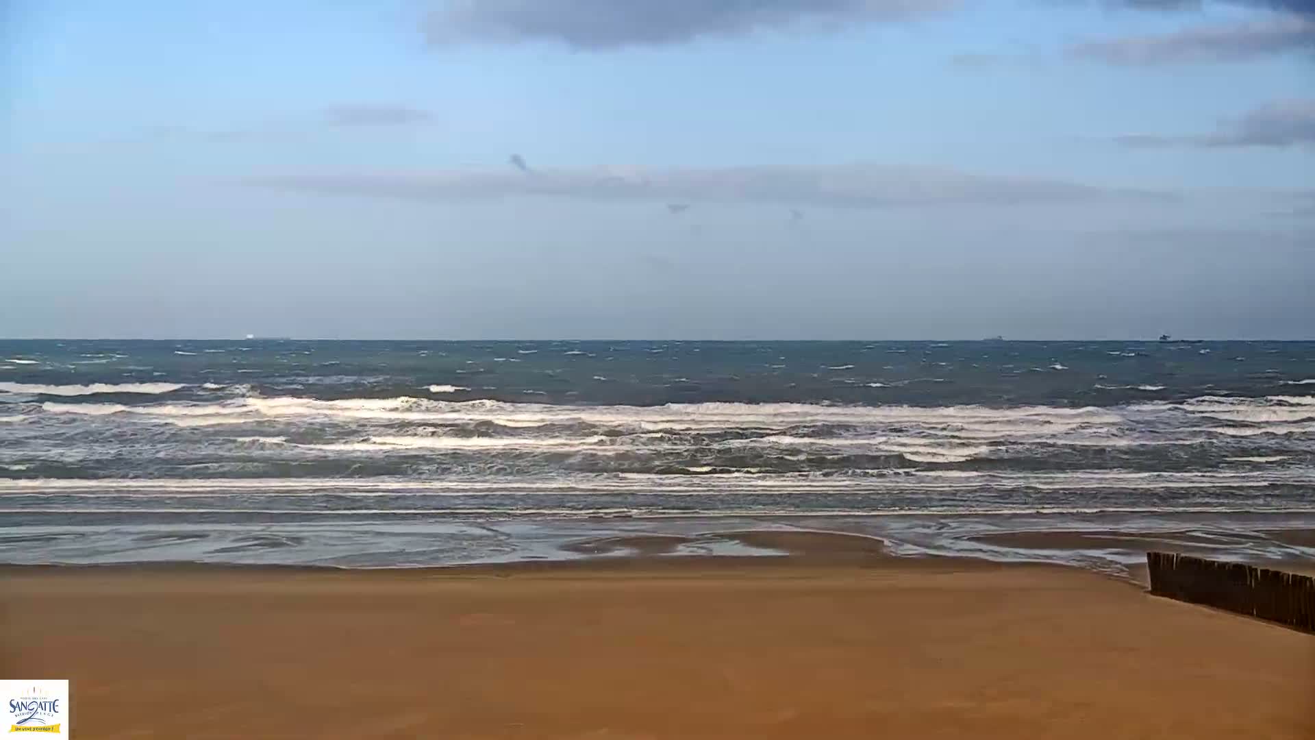 A windswept sandy beach with foamy waves rolling in from a choppy sea under a partly cloudy sky, with several ships visible on the distant horizon and a wooden groyne on the lower right.