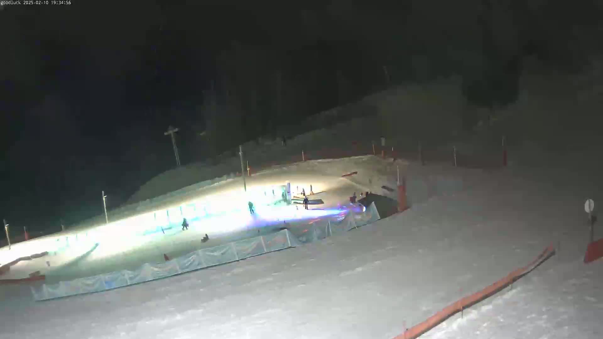 A brightly lit snow-covered area at night, featuring several people, appears to be a snow tubing or sledding hill.