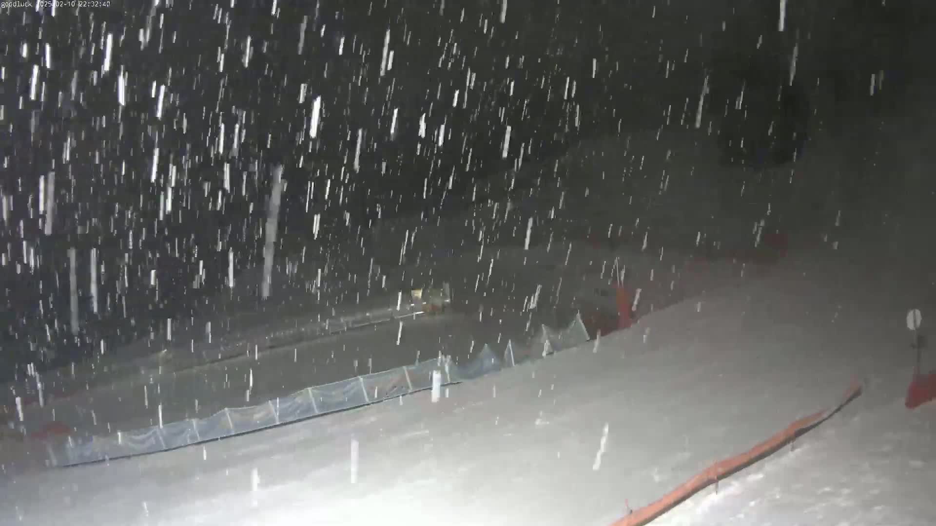 Heavy snowfall at night blankets a snow-covered slope with a low fence and orange structures.