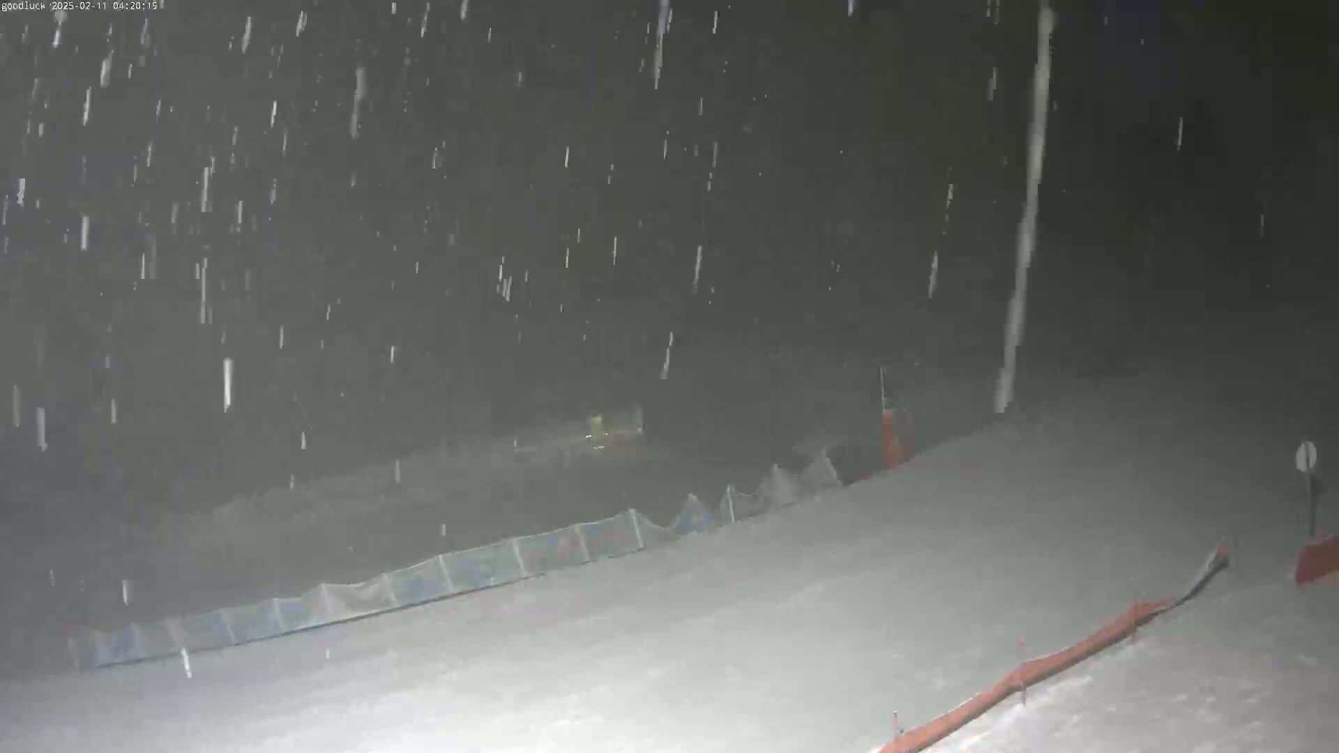 A snowy nighttime scene shows a snow-covered slope with a fence and some structures in the background under heavy snowfall.