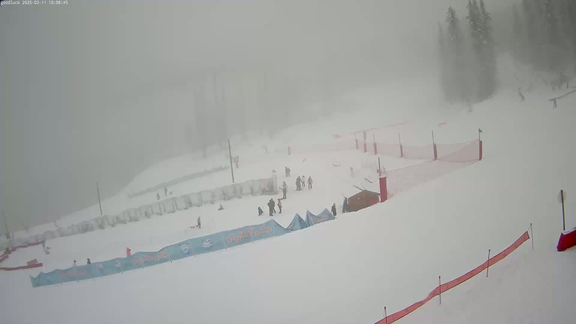 A snowy ski slope with several skiers and snowboarders is visible through a heavy fog.