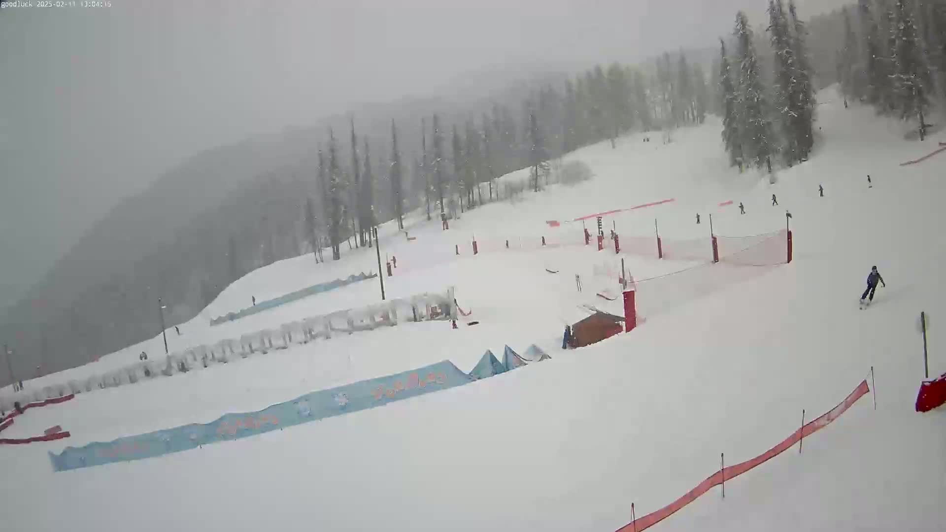 A snowy ski slope with several skiers and snowboarders visible, under heavy snowfall.