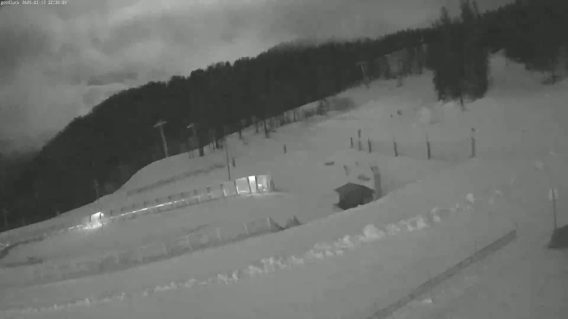 A nighttime, snow-covered landscape shows a mostly-empty ski slope with a small structure and a line of trees in the background under a cloudy sky.
