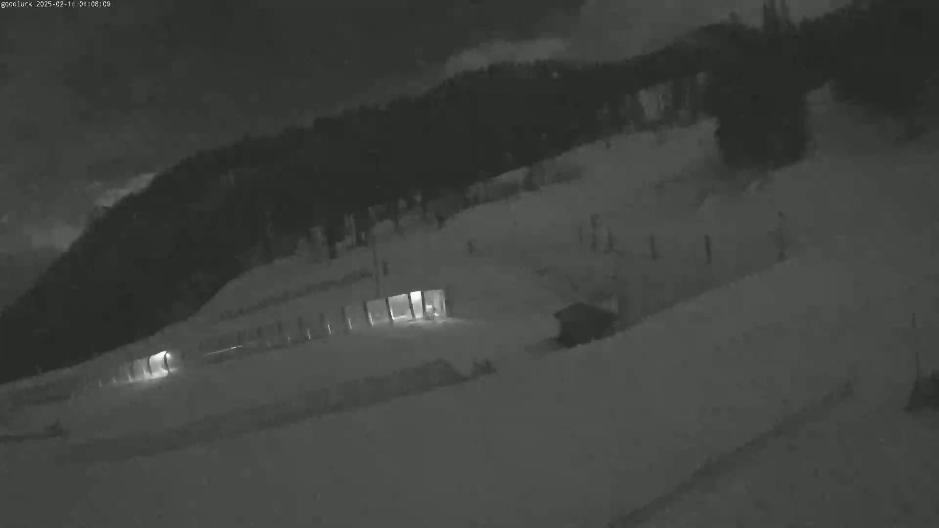 A nighttime, snow-covered landscape shows a building with lights on, illuminated against a dark, wooded hillside under a cloudy sky.