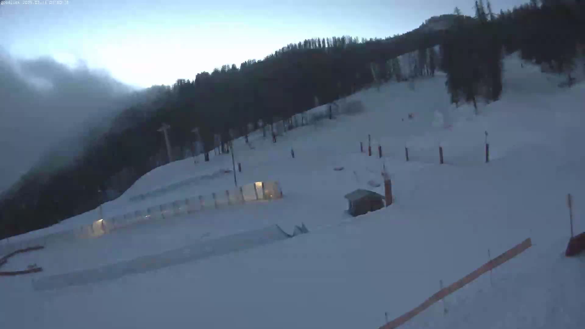 A snow-covered ski slope with a few structures and trees in the background under a cloudy, dusky sky.