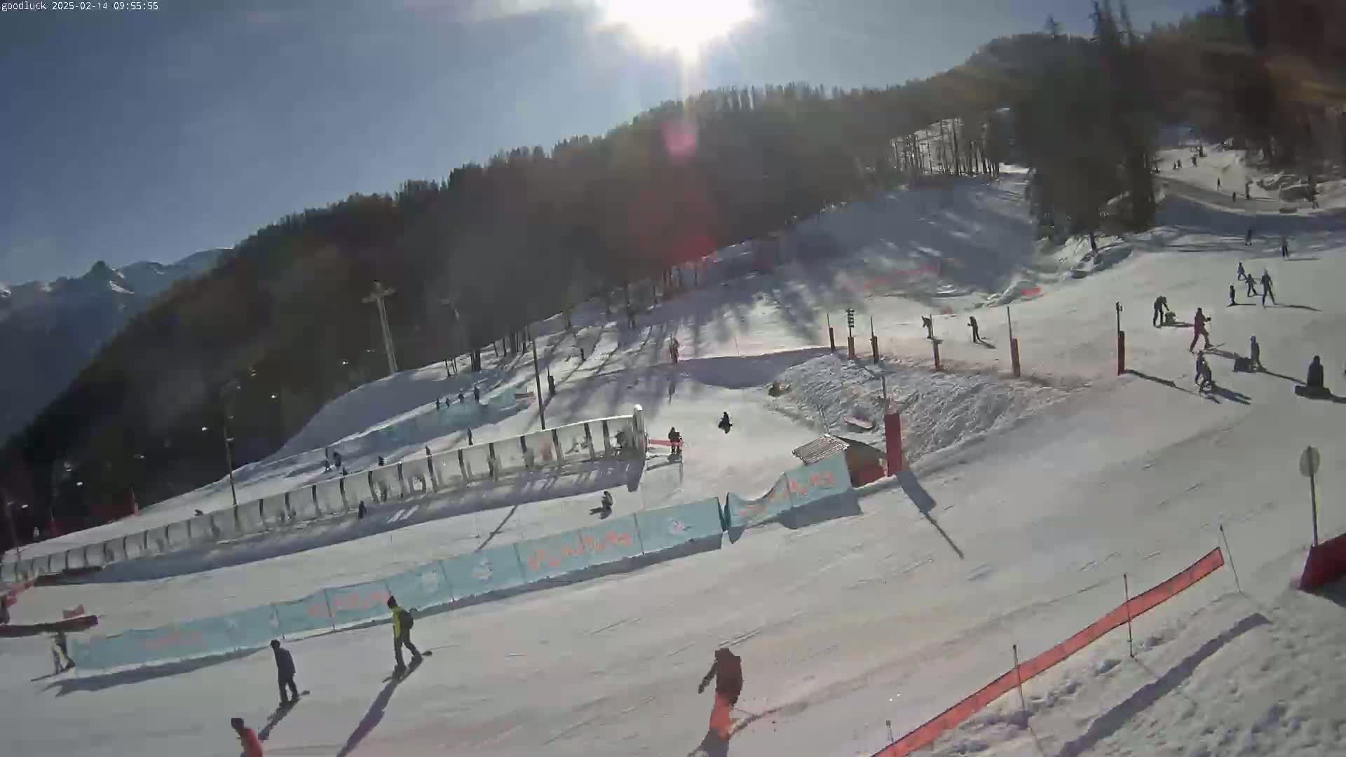 A sunny day at a snow-covered ski slope shows several people skiing and snowboarding down the hill, with mountains visible in the background.