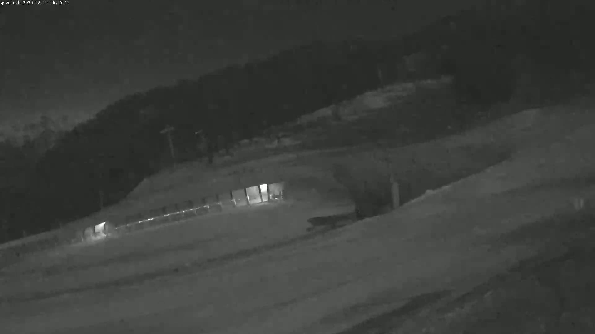 A nighttime, snowy scene shows a structure with lights on, a snow-covered slope, and a dark, treeline in the background.