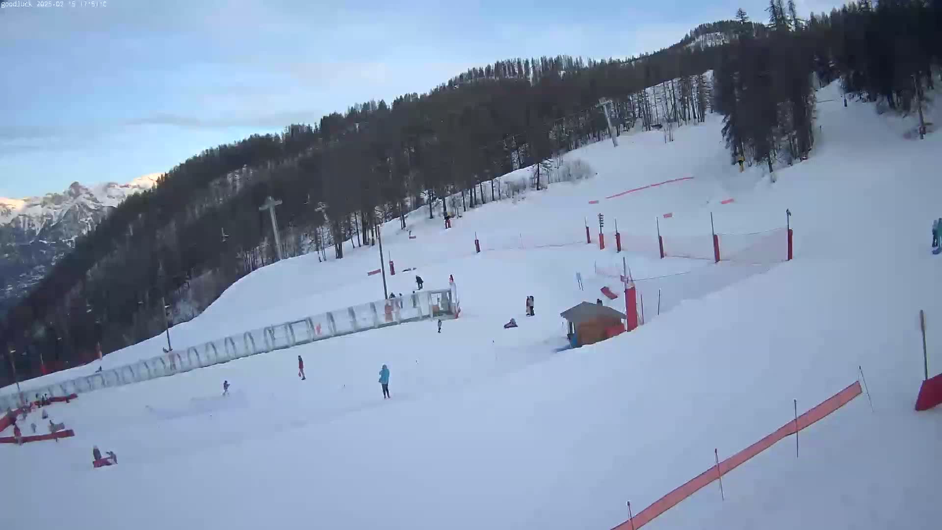 A snow-covered ski slope with several skiers and snowboarders, a small building, and safety netting, under a mostly clear sky.