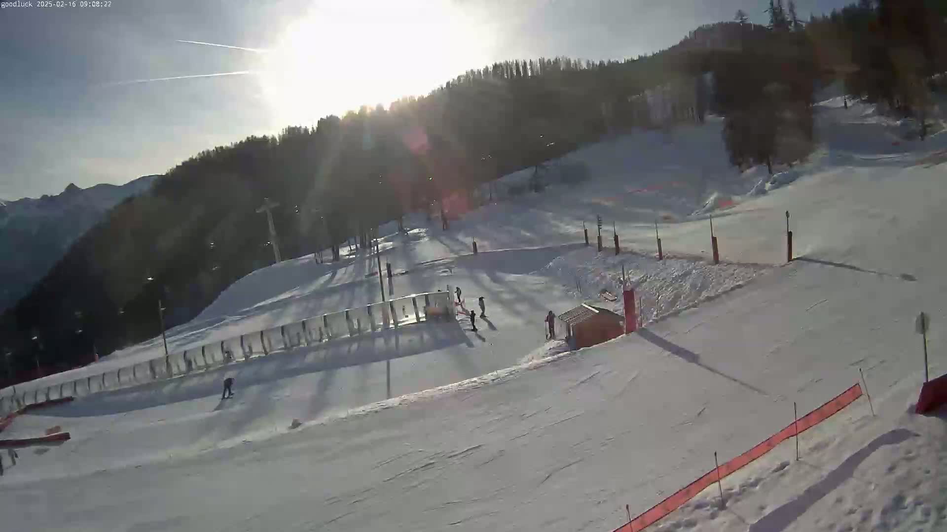 A sunny day on a snow-covered ski slope with several skiers and snowboarders visible, a ski lift in the background, and trees lining the hill.