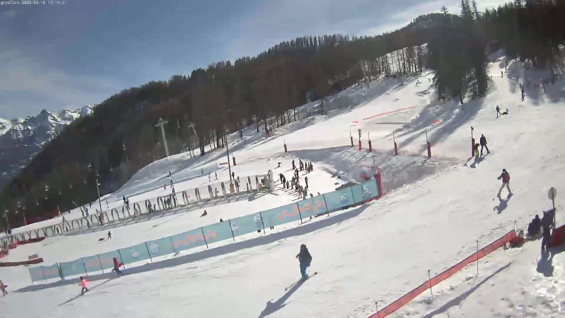 A snowy ski slope with several skiers and snowboarders under a sunny sky, mountains are visible in the background.