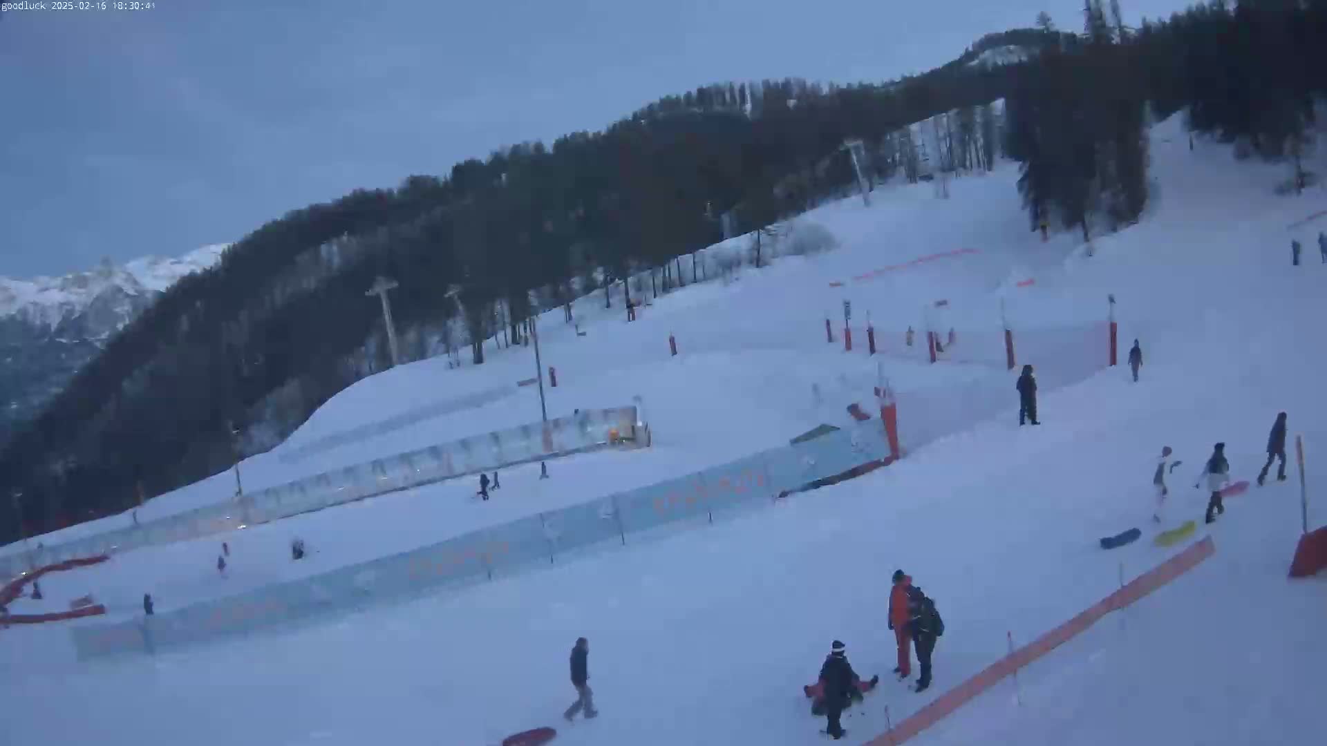A snow-covered ski slope with several people skiing, snowboarding, and walking, under an overcast sky.