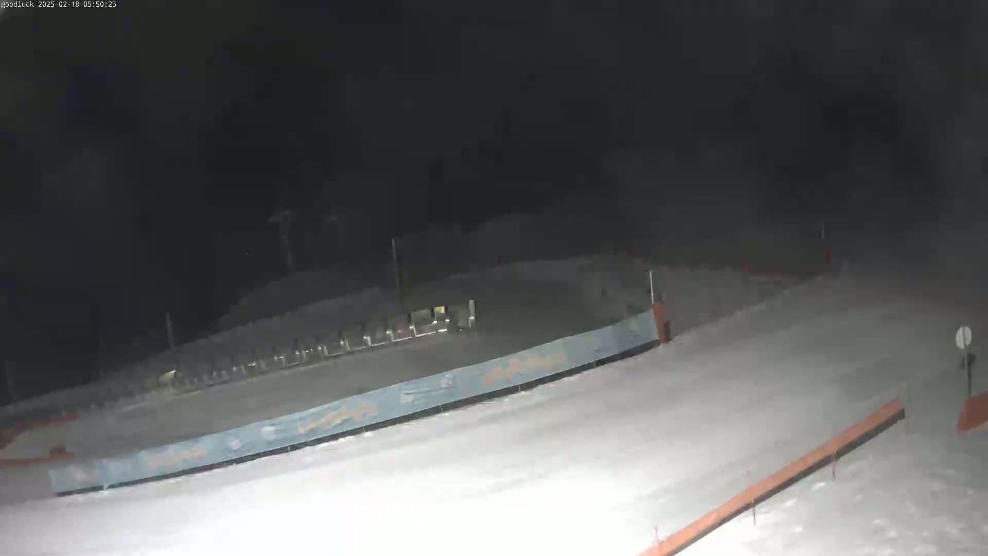 A snow-covered slope at night is partially illuminated, with a fence and some structures visible in the distance.