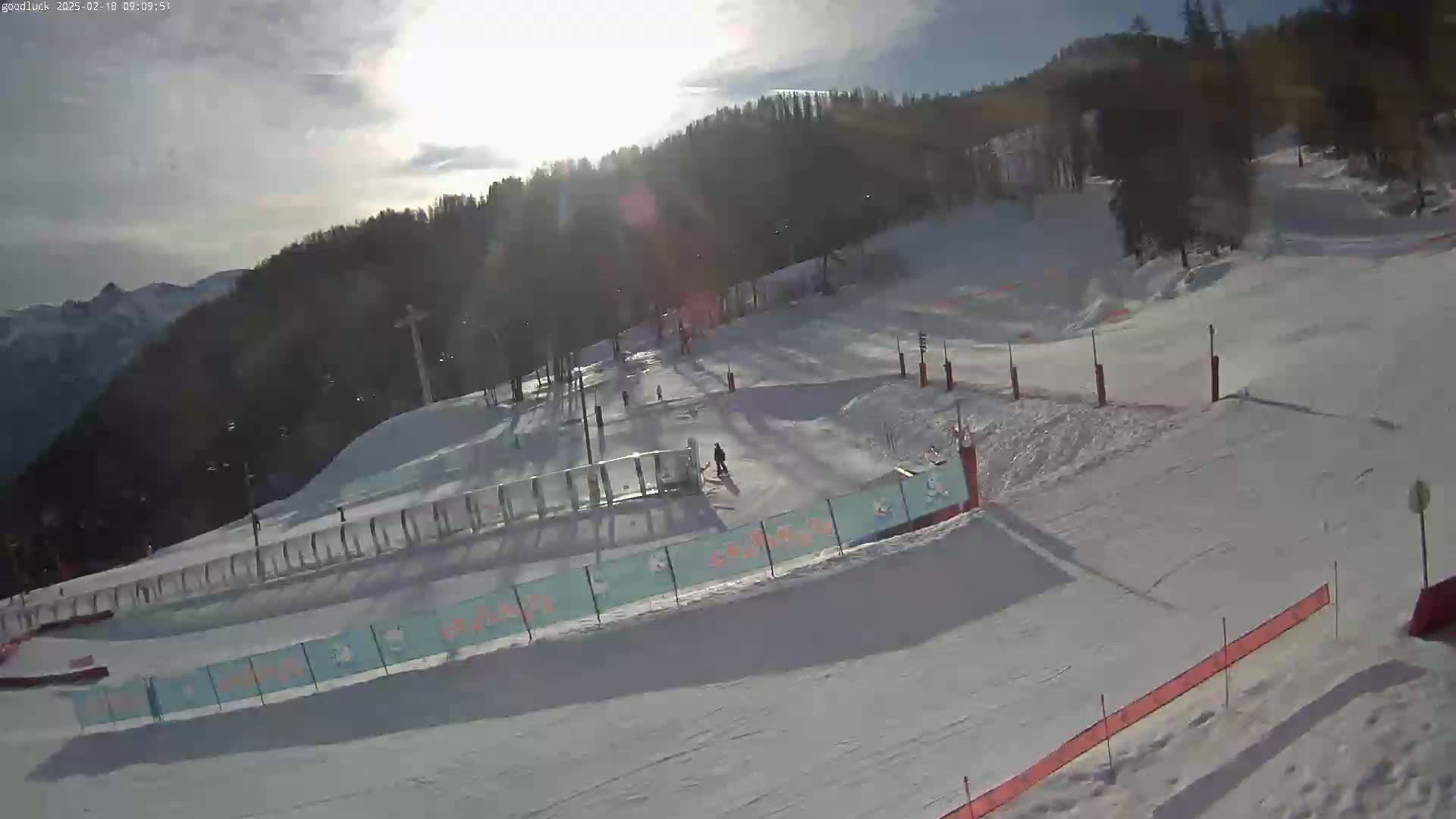 A sunny day at a snow-covered ski slope shows several skiers and snowboarders on the runs, with trees and mountains in the background.