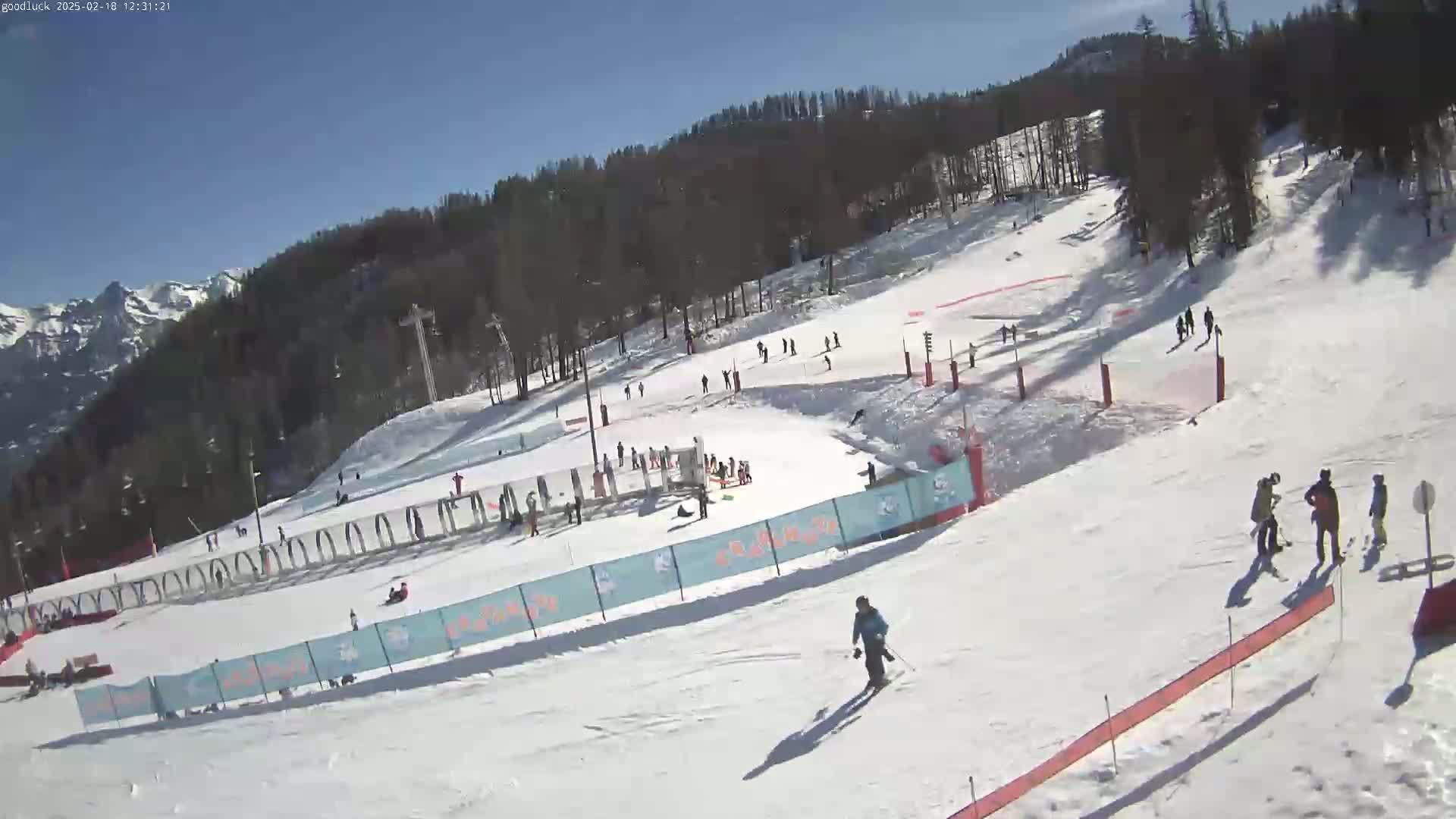 A sunny day at a snow-covered ski resort shows several skiers and snowboarders on the slopes, with trees and mountains in the background.