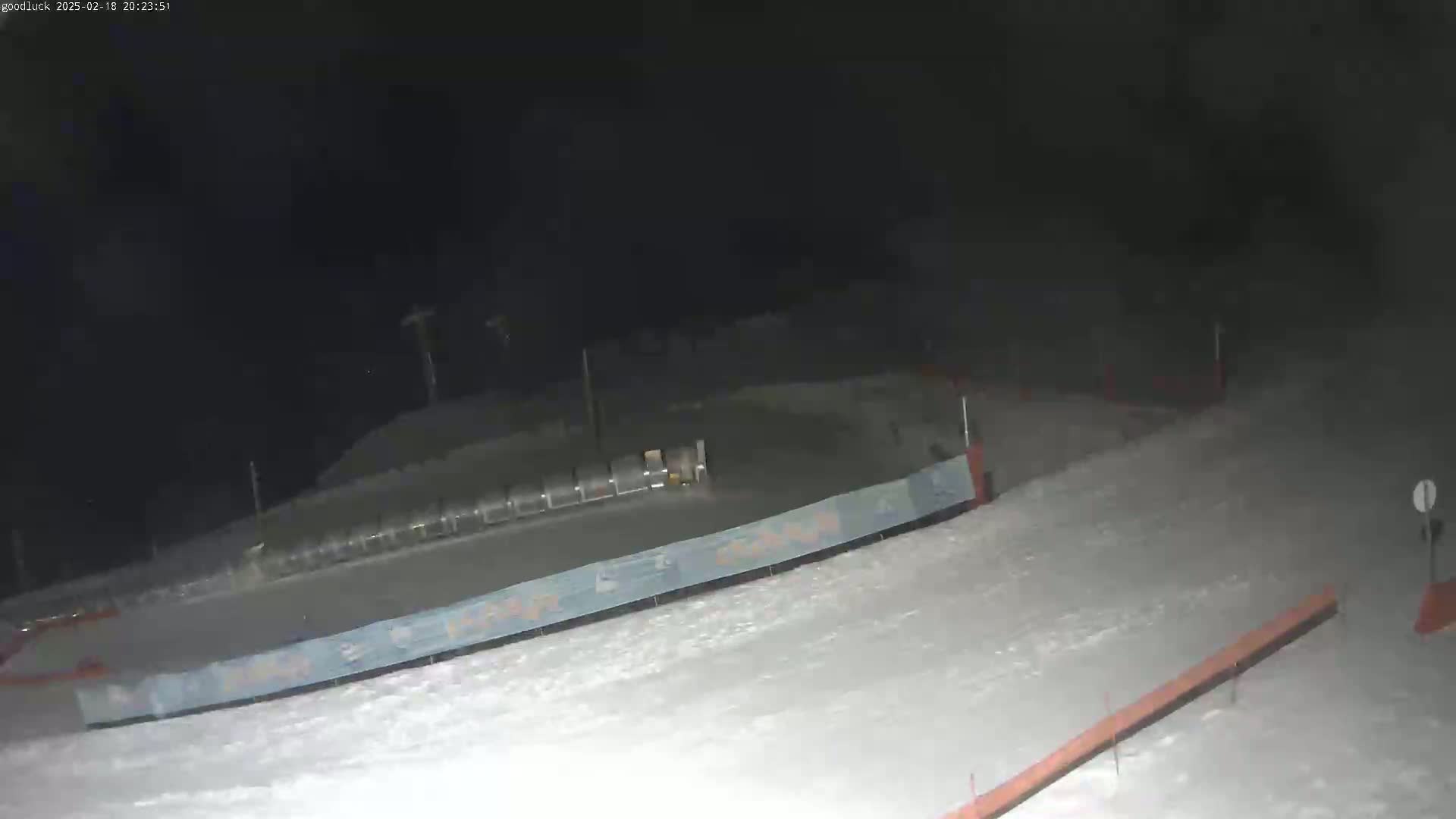 A nighttime view of a snow-covered slope with a barrier and what appears to be a ski lift in the background.