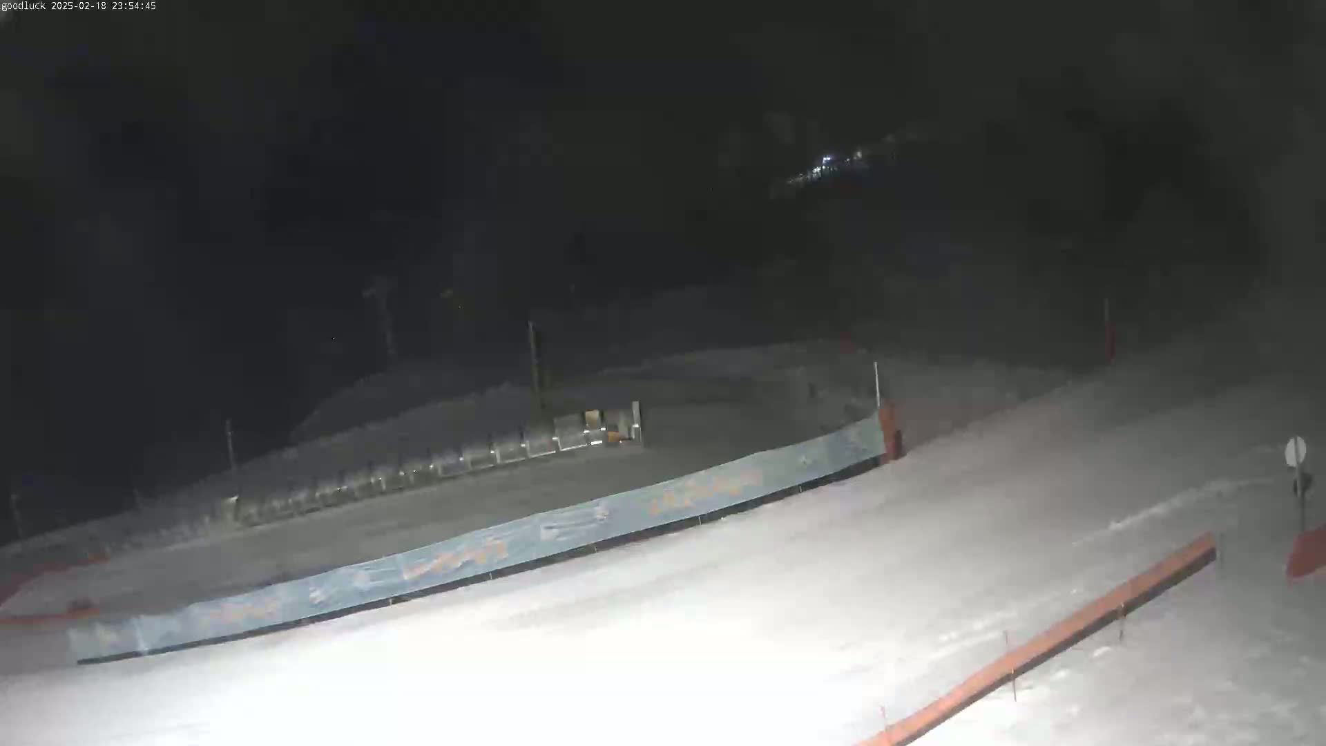A snow-covered ski slope is visible at night, with a long, low barrier and some other structures in the background.