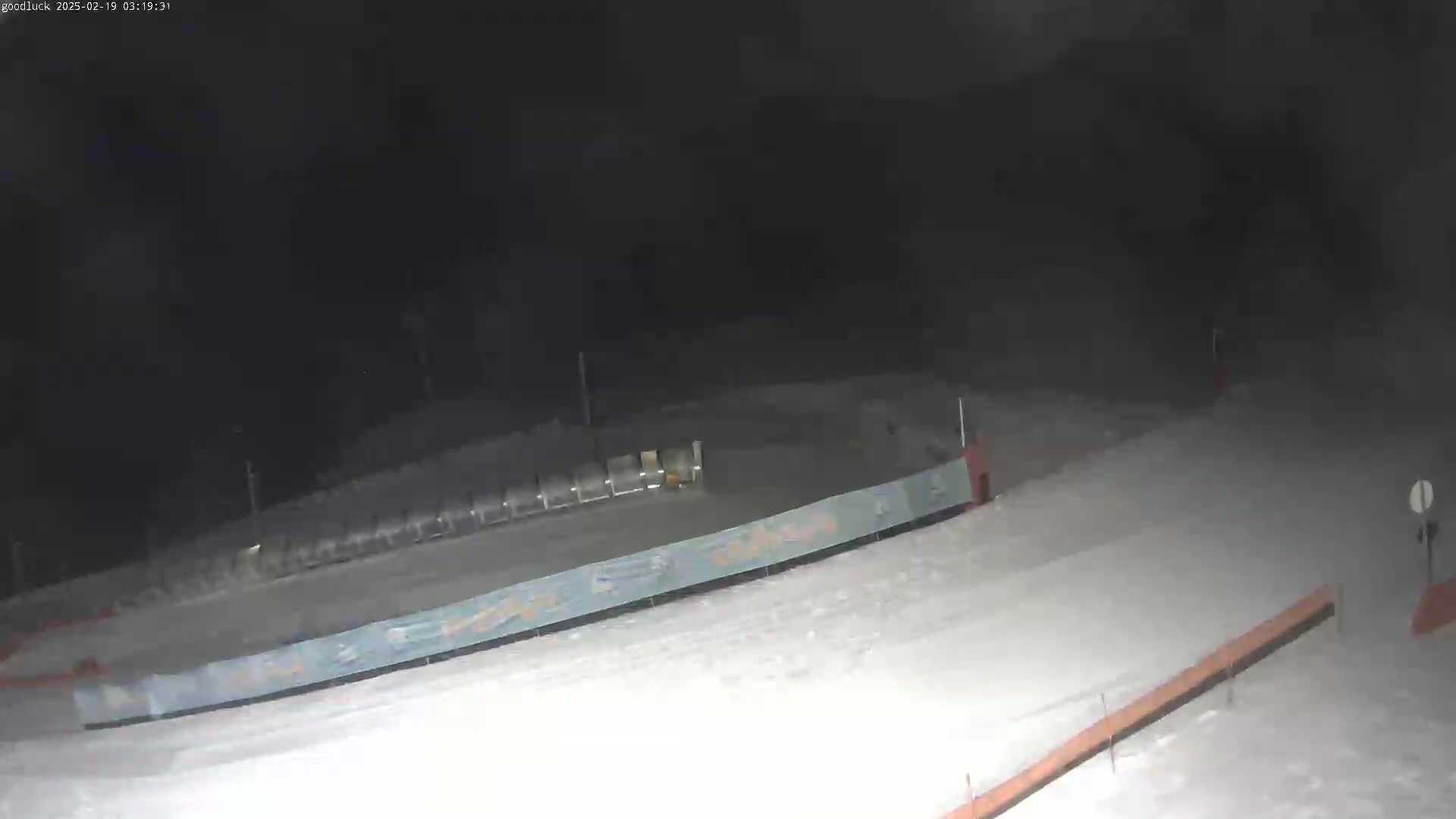 A snow-covered slope at night, with a lift and fencing visible under dark skies.