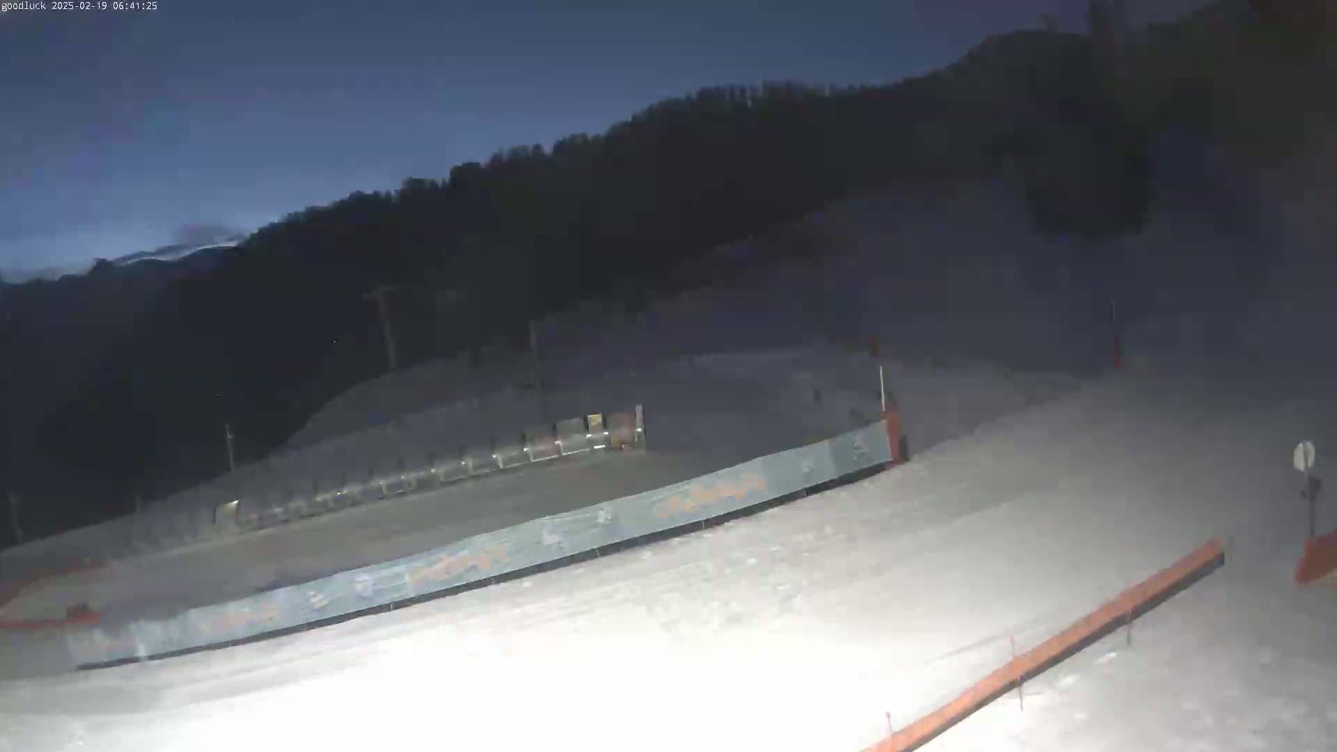 A snow-covered slope at night is dimly lit, with a dark mountain in the background under a twilight sky.