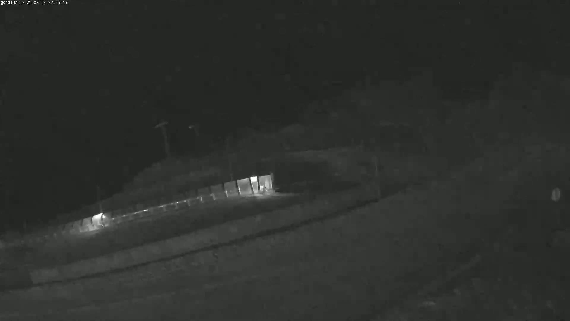 A nighttime, low-light security camera image shows a dimly lit area with a fence and a structure, possibly a garage or shed, with an illuminated interior.