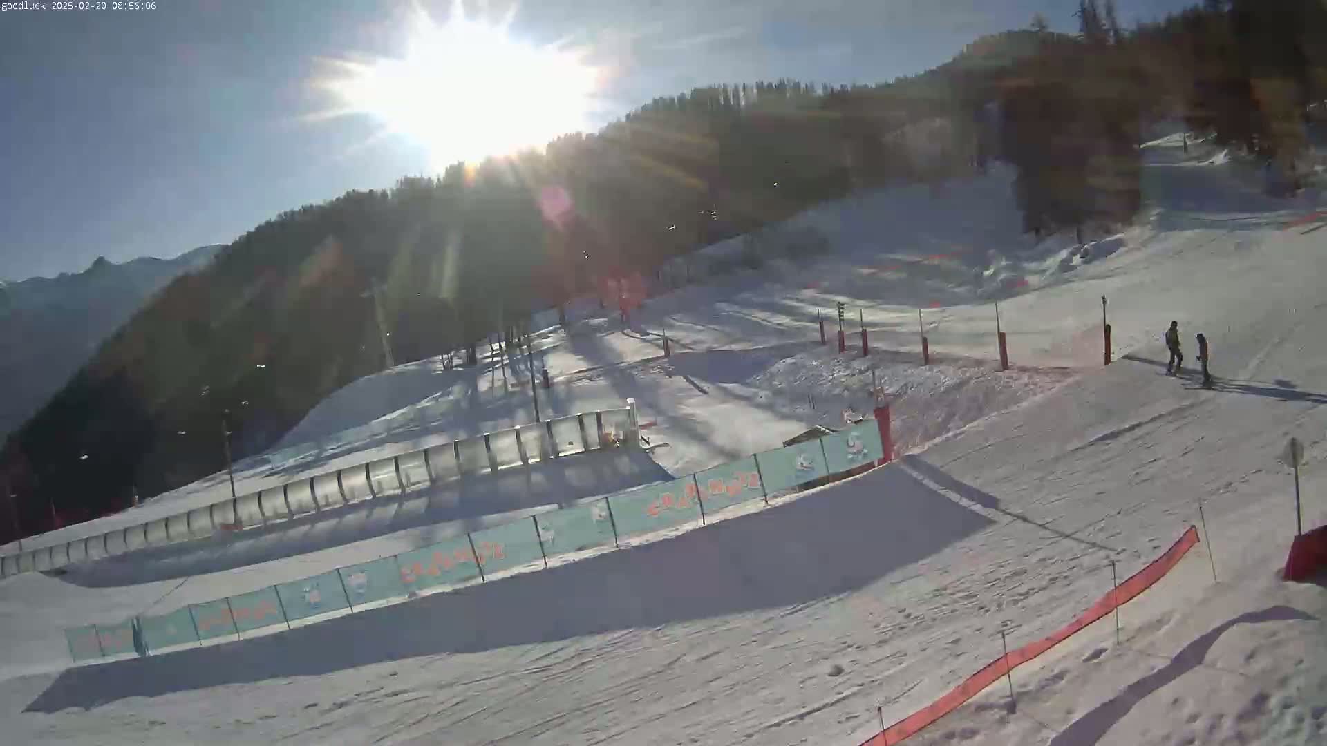 A sunny, snow-covered ski slope with two people walking near a fence and a mountain in the background.