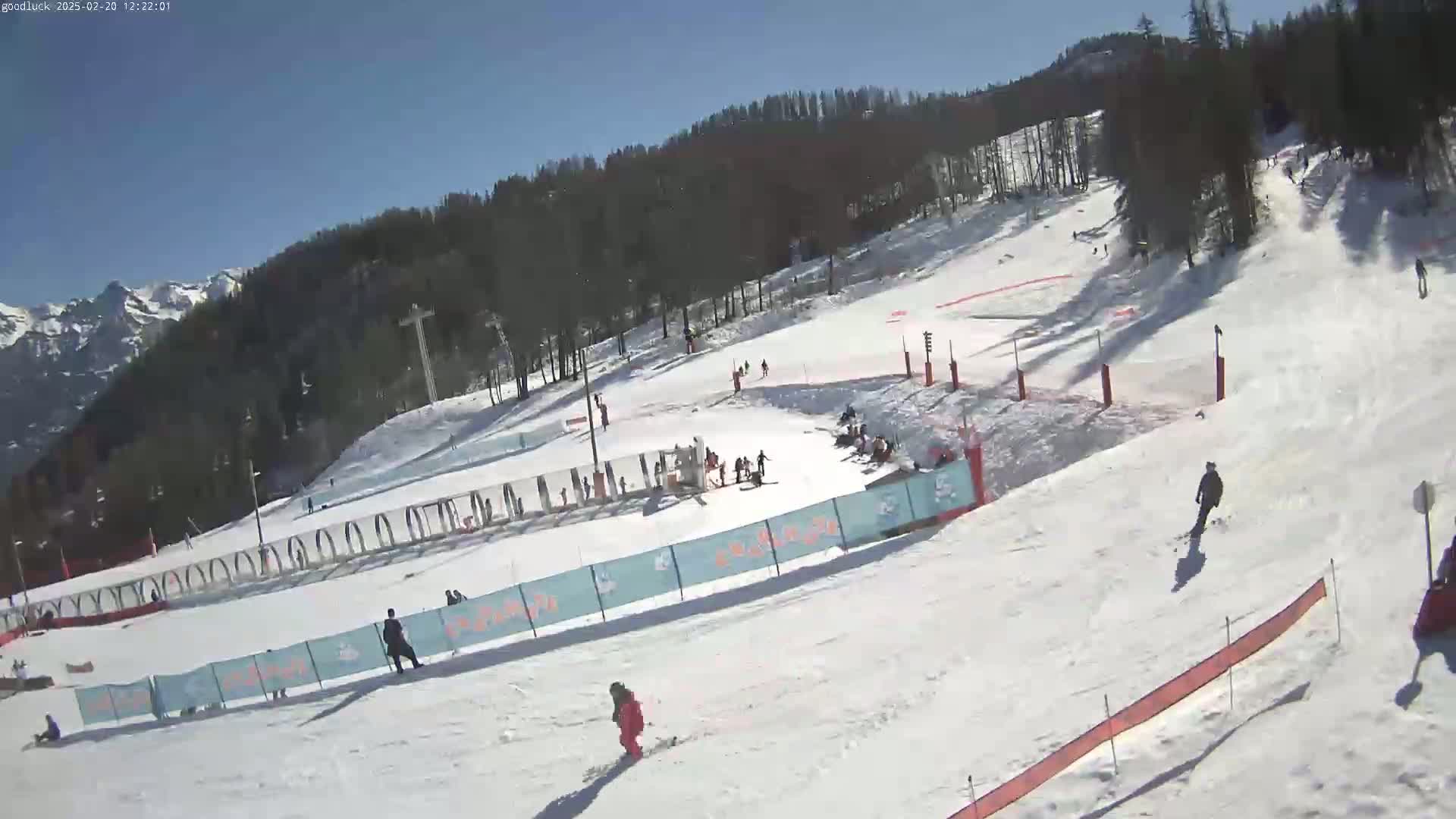A sunny day at a snow-covered ski slope shows several skiers and snowboarders enjoying the mountainside, with trees and distant mountains in the background.