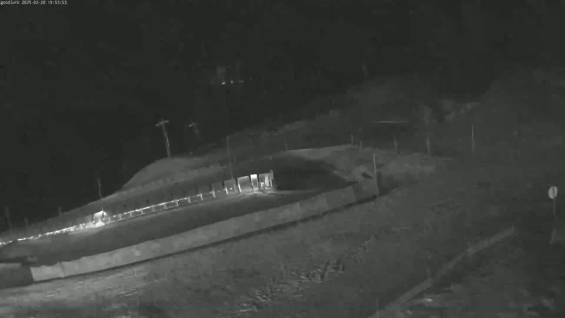 A nighttime, low-light image shows a fenced-in area with what appears to be a road or path leading to it, under a dark, clear sky.