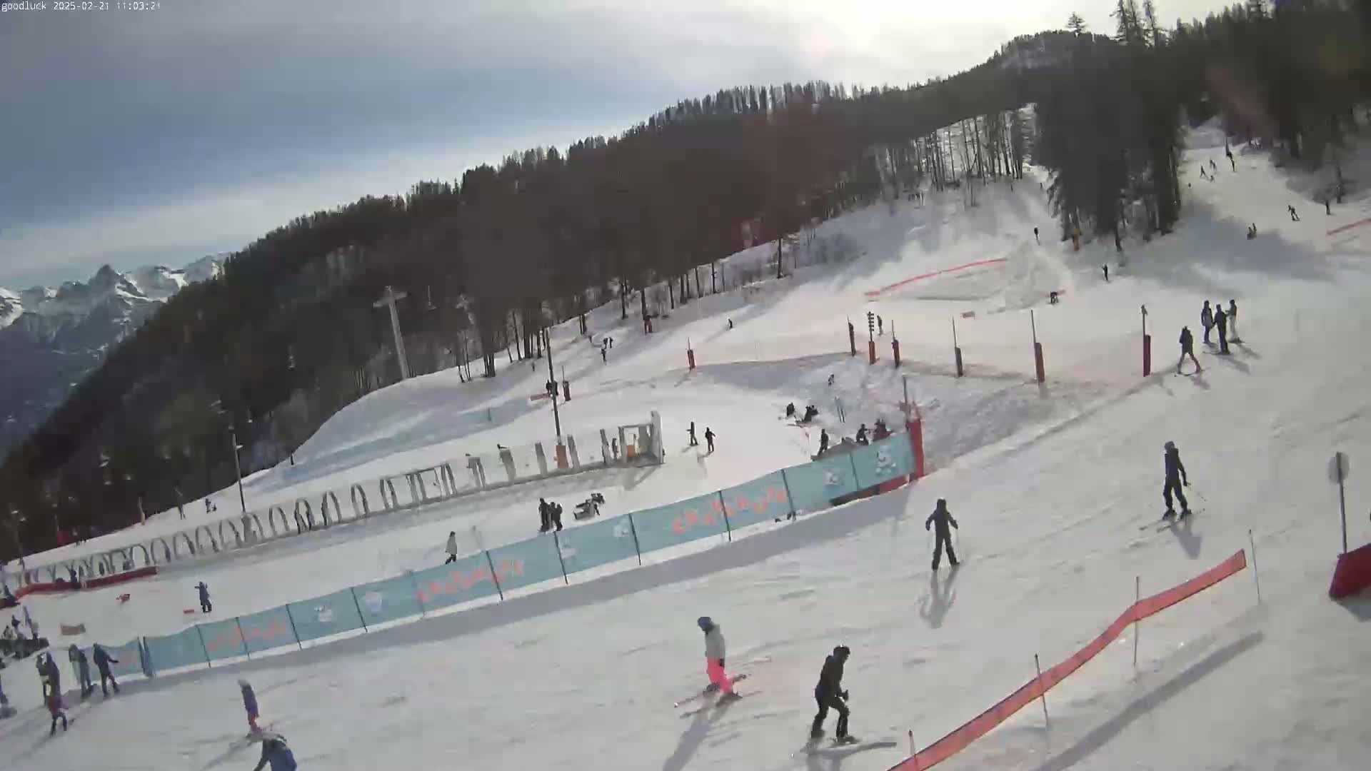 A snowy ski slope with several skiers and snowboarders, under a partly cloudy sky, is surrounded by a forested mountain range in the background.