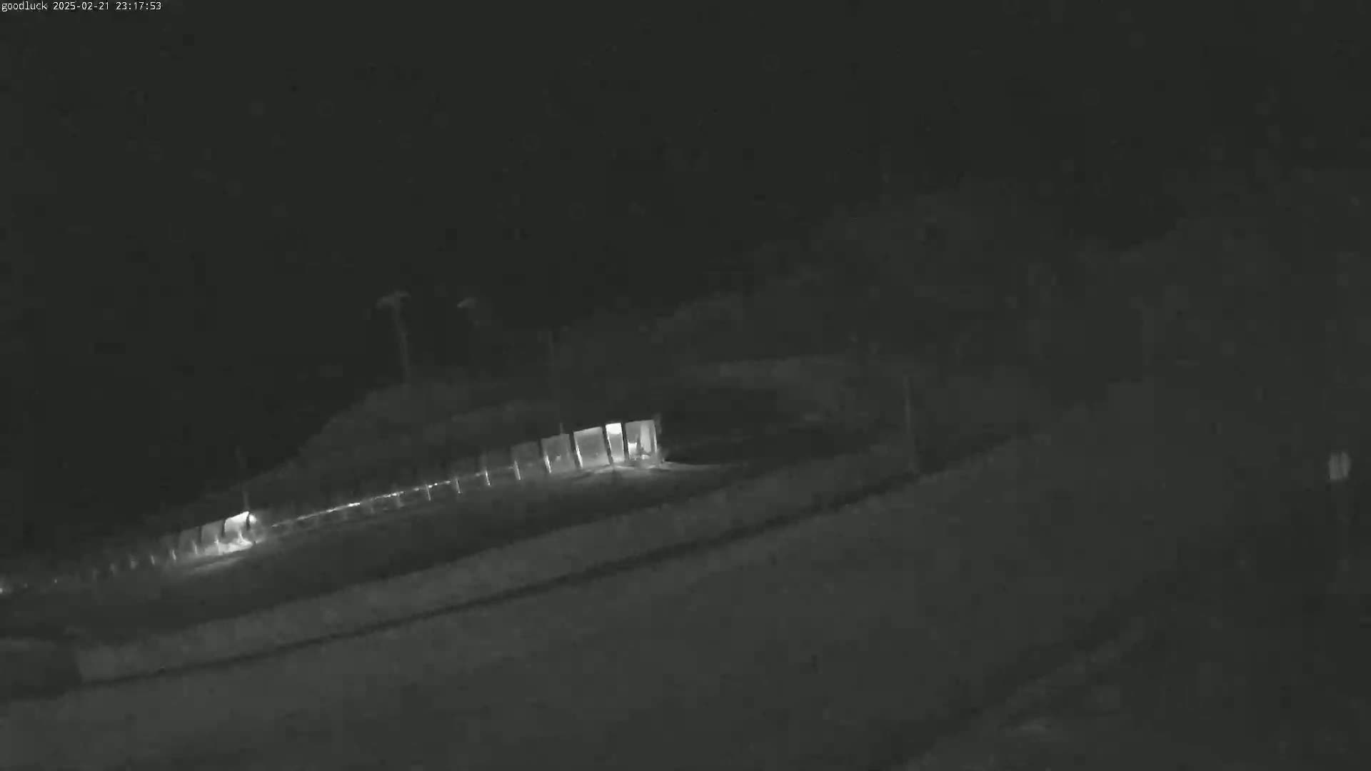 A nighttime, low-light image shows a dark roadway leading to a dimly lit structure under a dark sky.