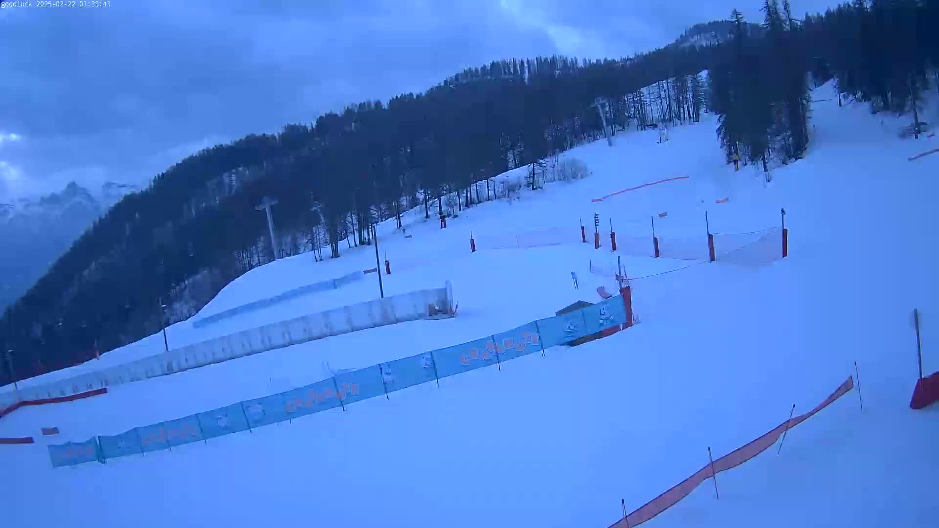 A snow-covered ski slope with several marked runs and safety nets is visible under an overcast sky.