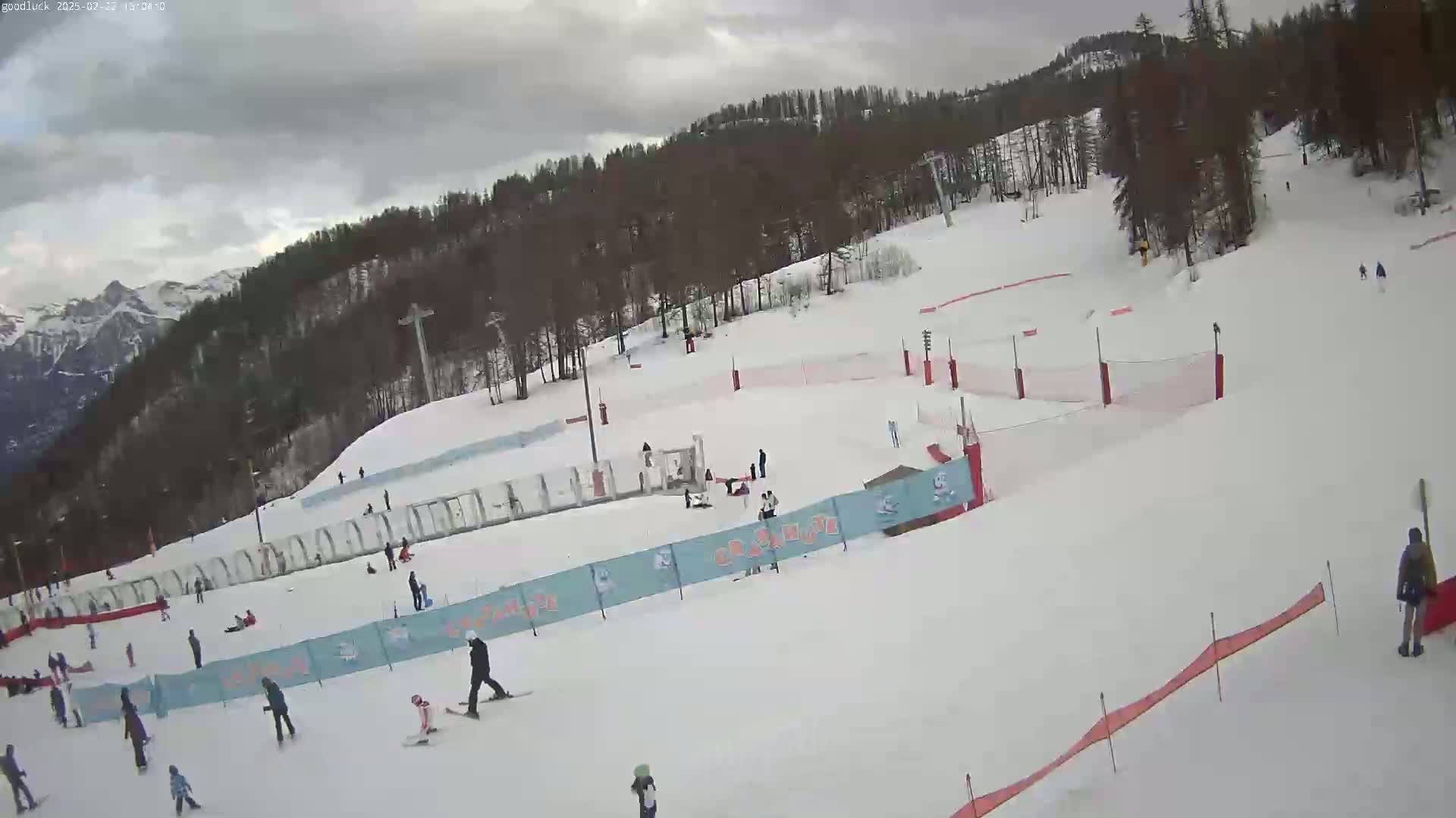 A snowy ski slope with several skiers and snowboarders, surrounded by trees and mountains under a cloudy sky.