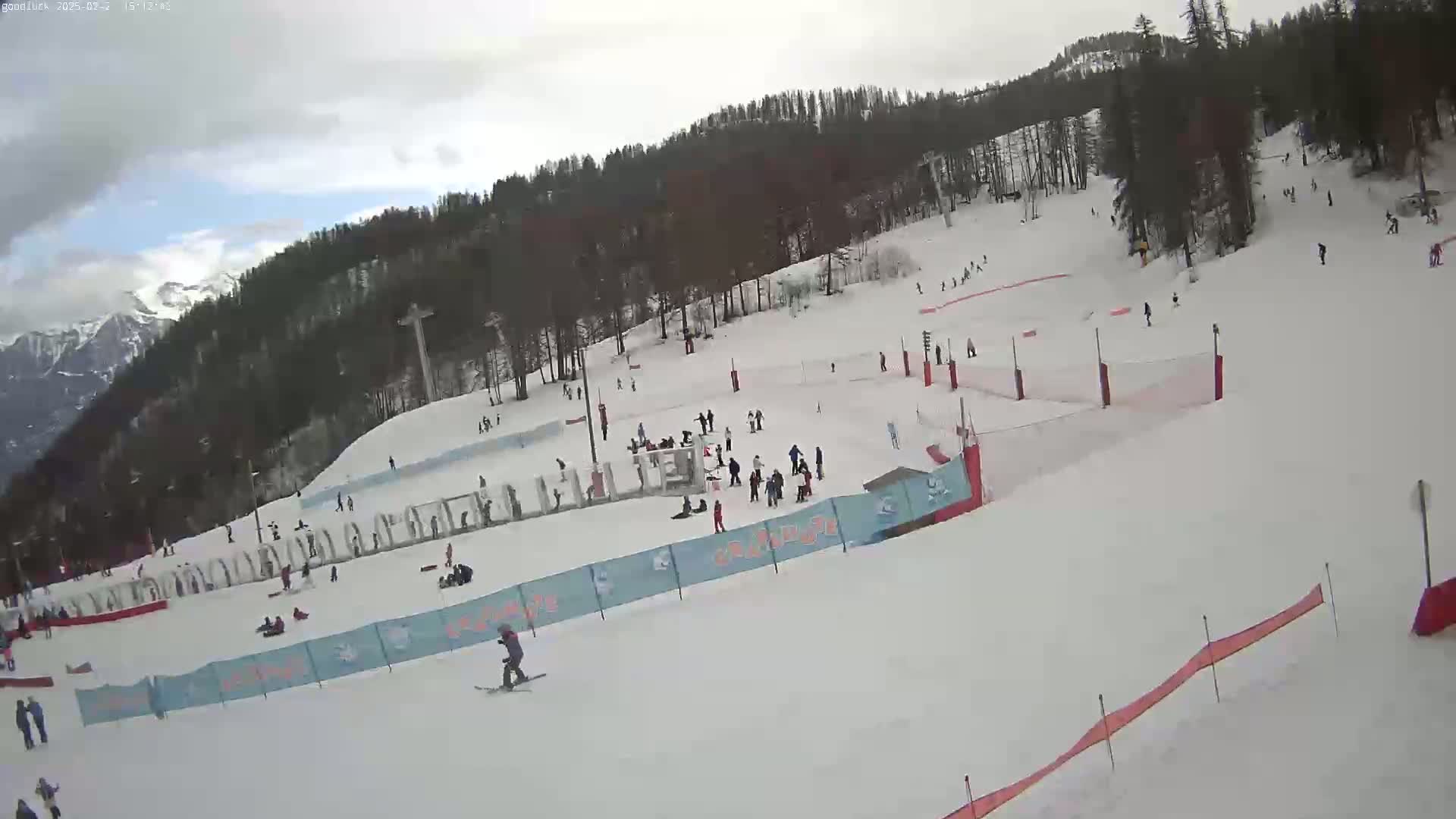 A snowy ski slope with several skiers and snowboarders, surrounded by a forest and mountains under a cloudy sky.