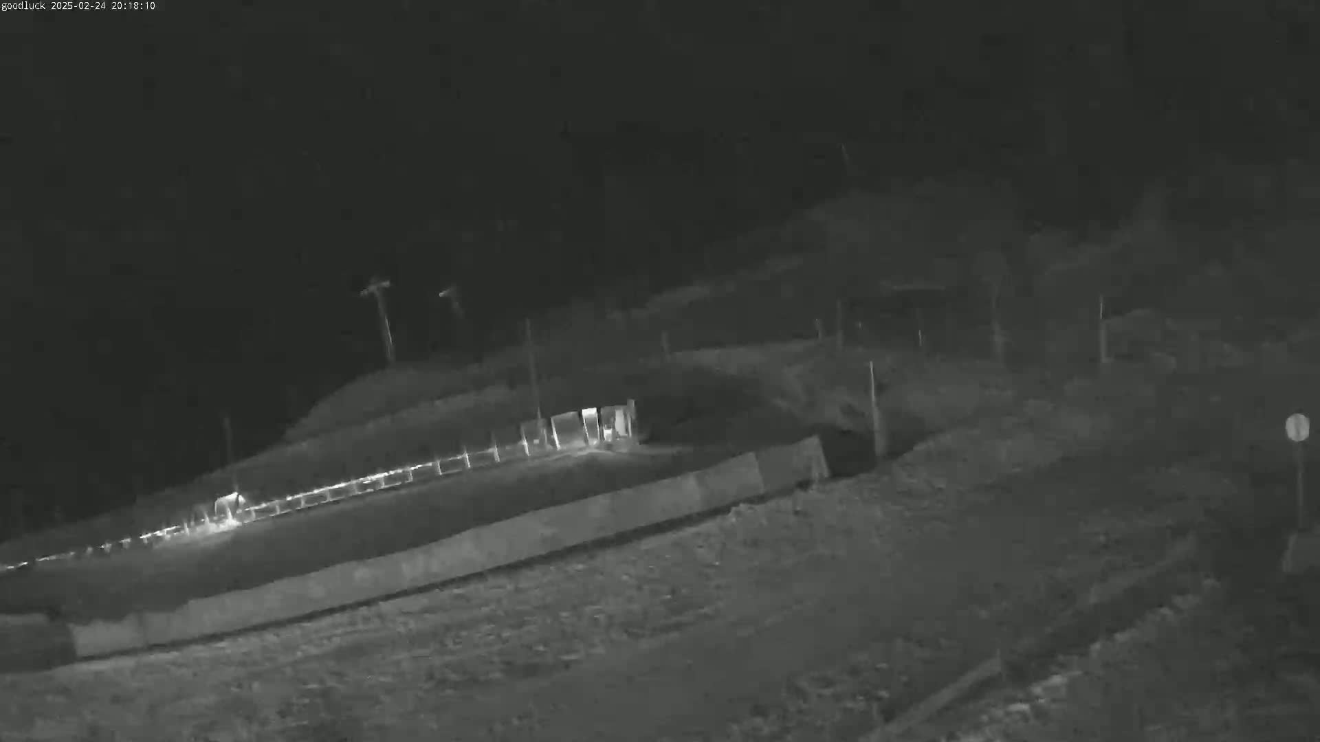 A nighttime, low-light image shows a dark, unpaved area with a low wall and some distant lights.
