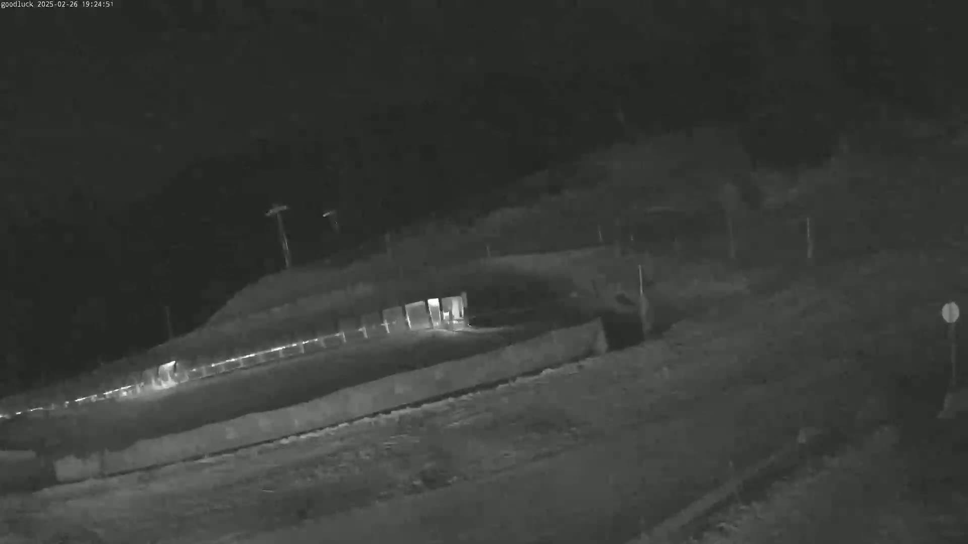 A nighttime, low-light image shows a roadway with a fence and some structures in the background.