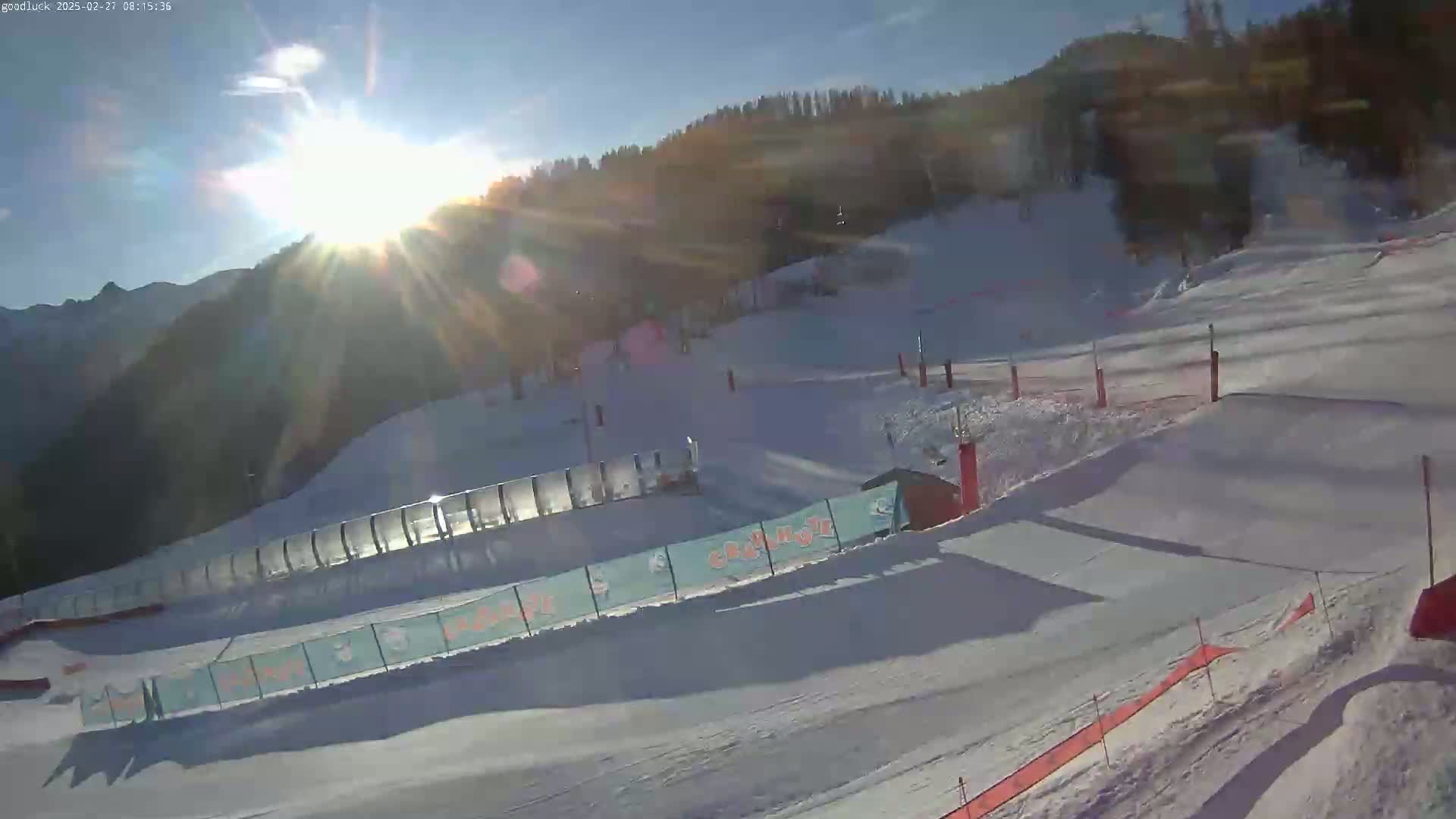 A sunny, snow-covered ski slope with various features including fences, ramps, and signage is visible, mountains in the background.