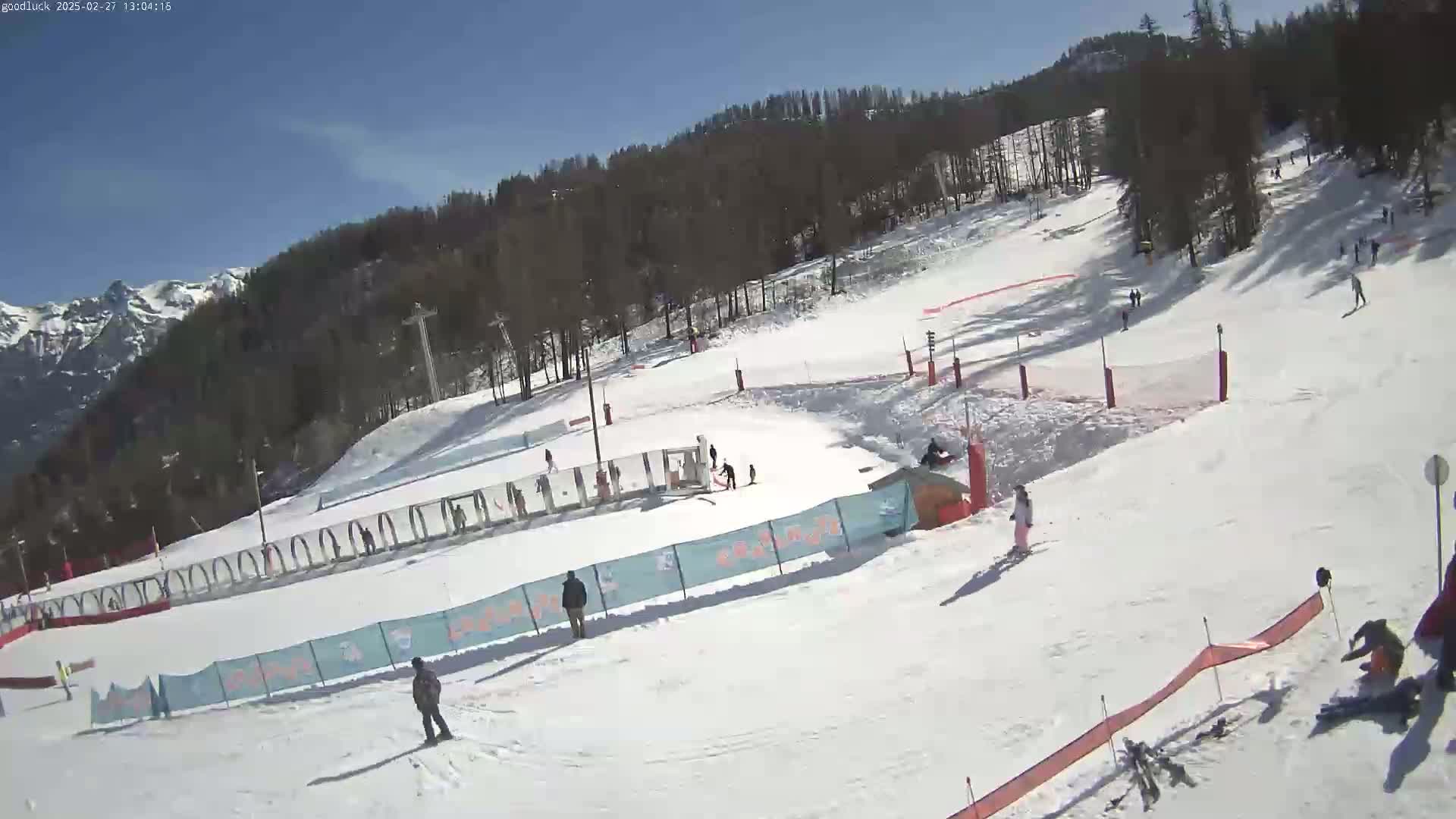 A sunny day on a snow-covered ski slope with several skiers and snowboarders present, surrounded by trees and mountains in the background.