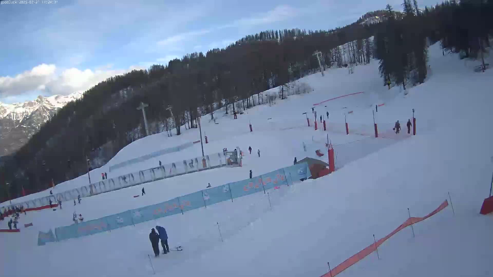 A mostly sunny day at a snow-covered ski resort shows several skiers and snowboarders on the slopes, with mountains in the background.