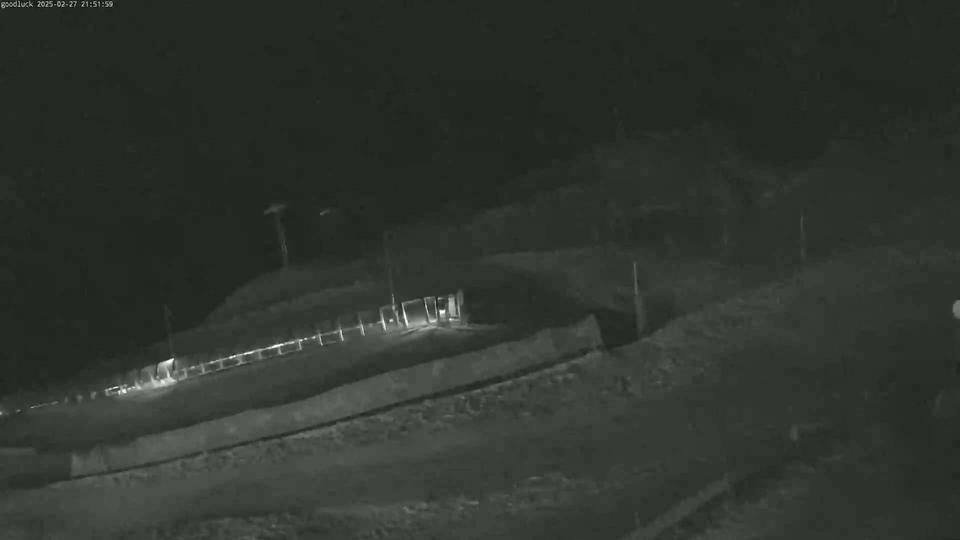 A nighttime, low-light view shows a dark, uneven landscape with a fence and some faint lights in the distance.