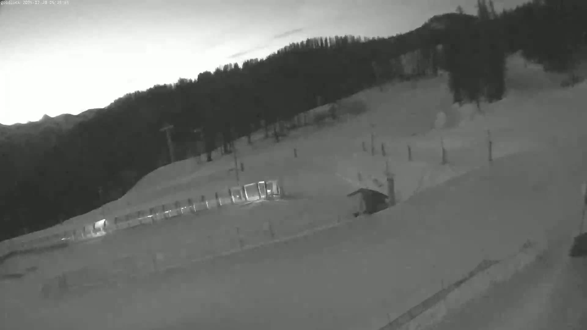 A nighttime, snow-covered ski slope with a ski lift and a few structures visible under a dusky sky.