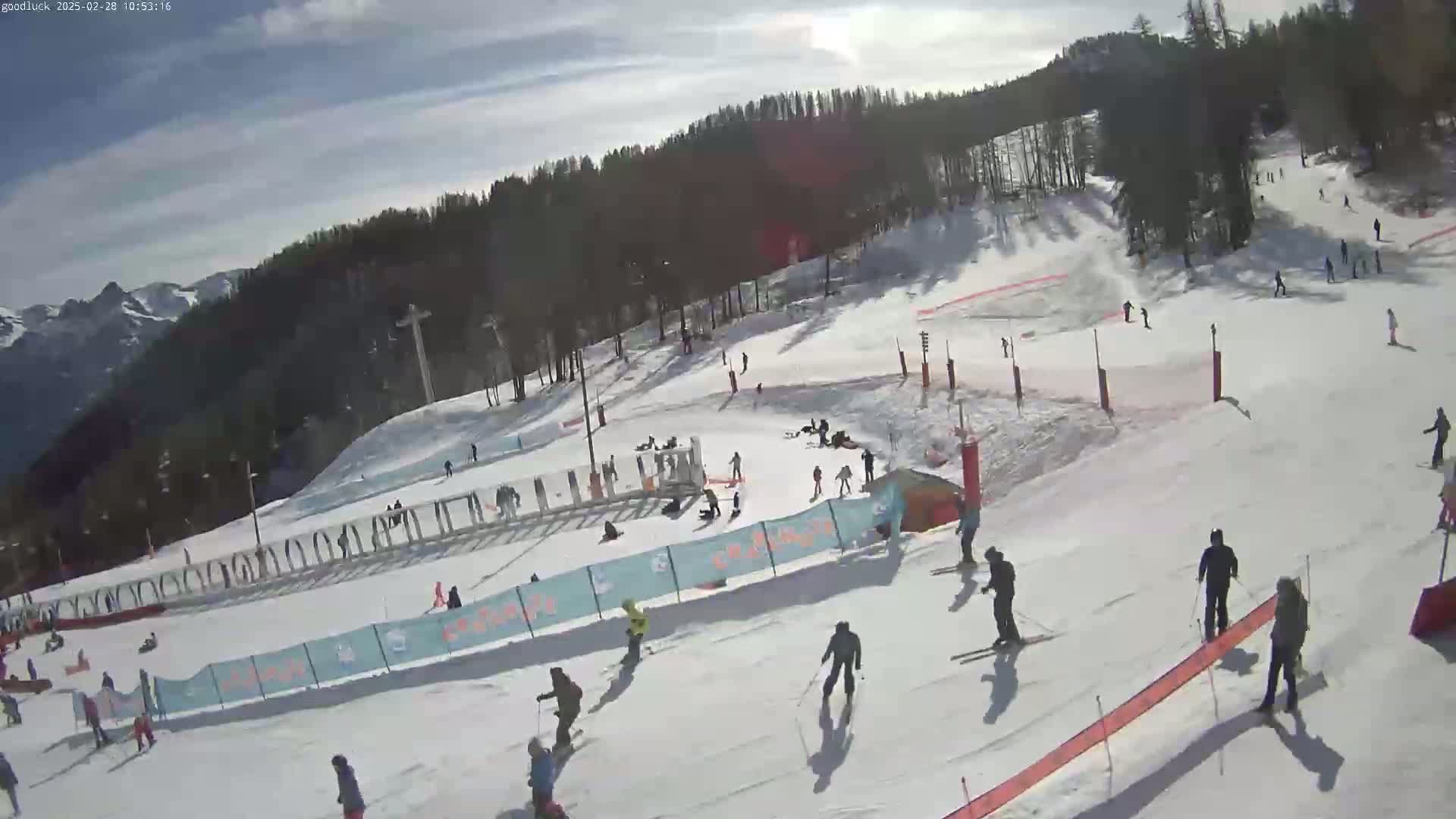 A snow-covered ski slope with numerous skiers and snowboarders, under a mostly sunny sky, is surrounded by a coniferous forest and distant mountains.