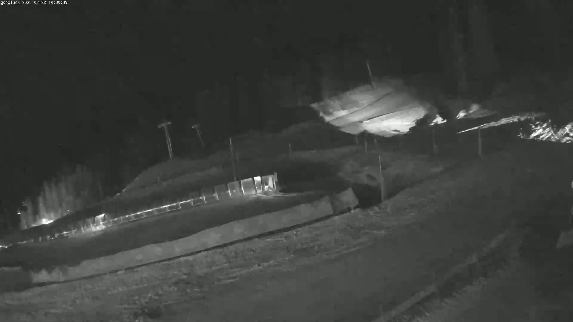 A nighttime, low-light image shows a sloped, snow-covered area with fencing and tall poles, possibly at a ski resort or similar location.