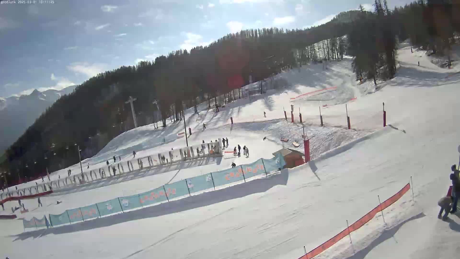 A snow-covered ski slope with several skiers and snowboarders, under a mostly sunny sky.