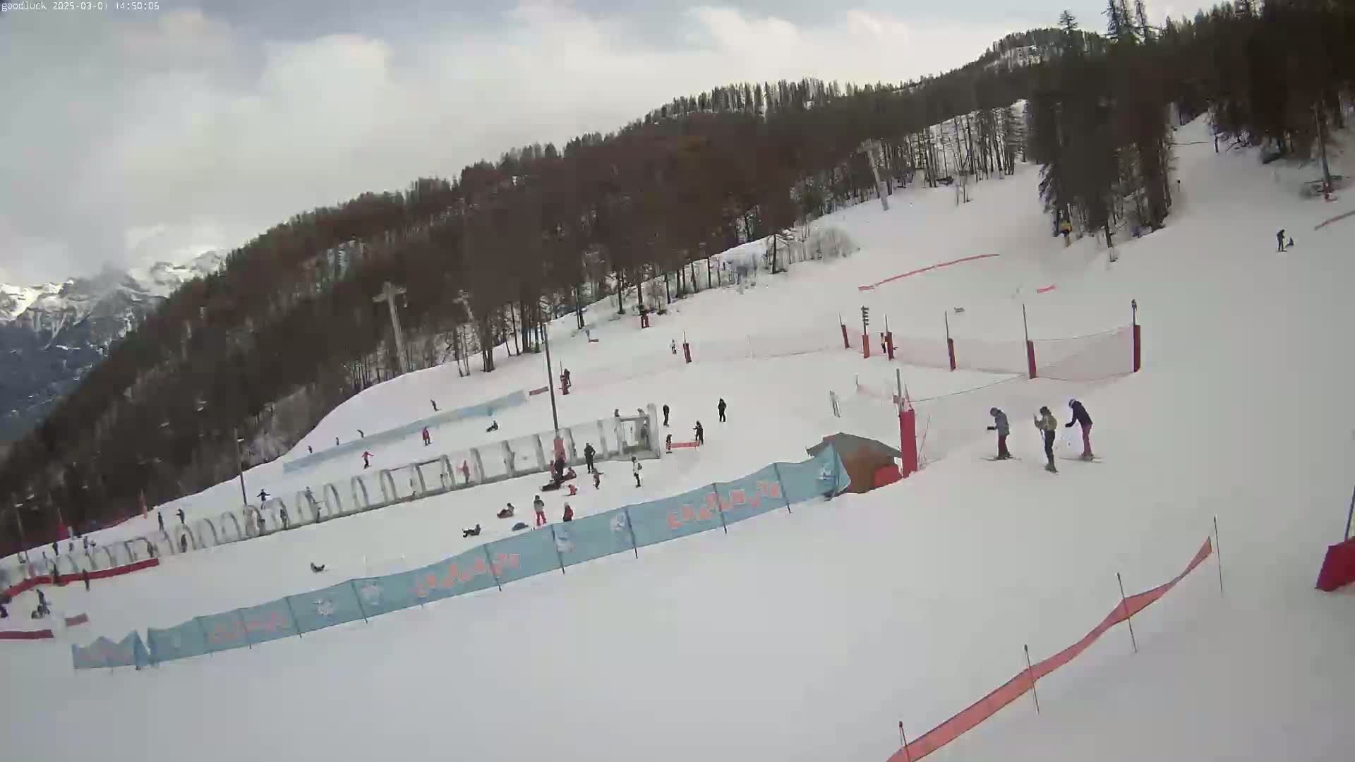 A snowy ski slope with several skiers and snowboarders, surrounded by trees and mountains under a cloudy sky.