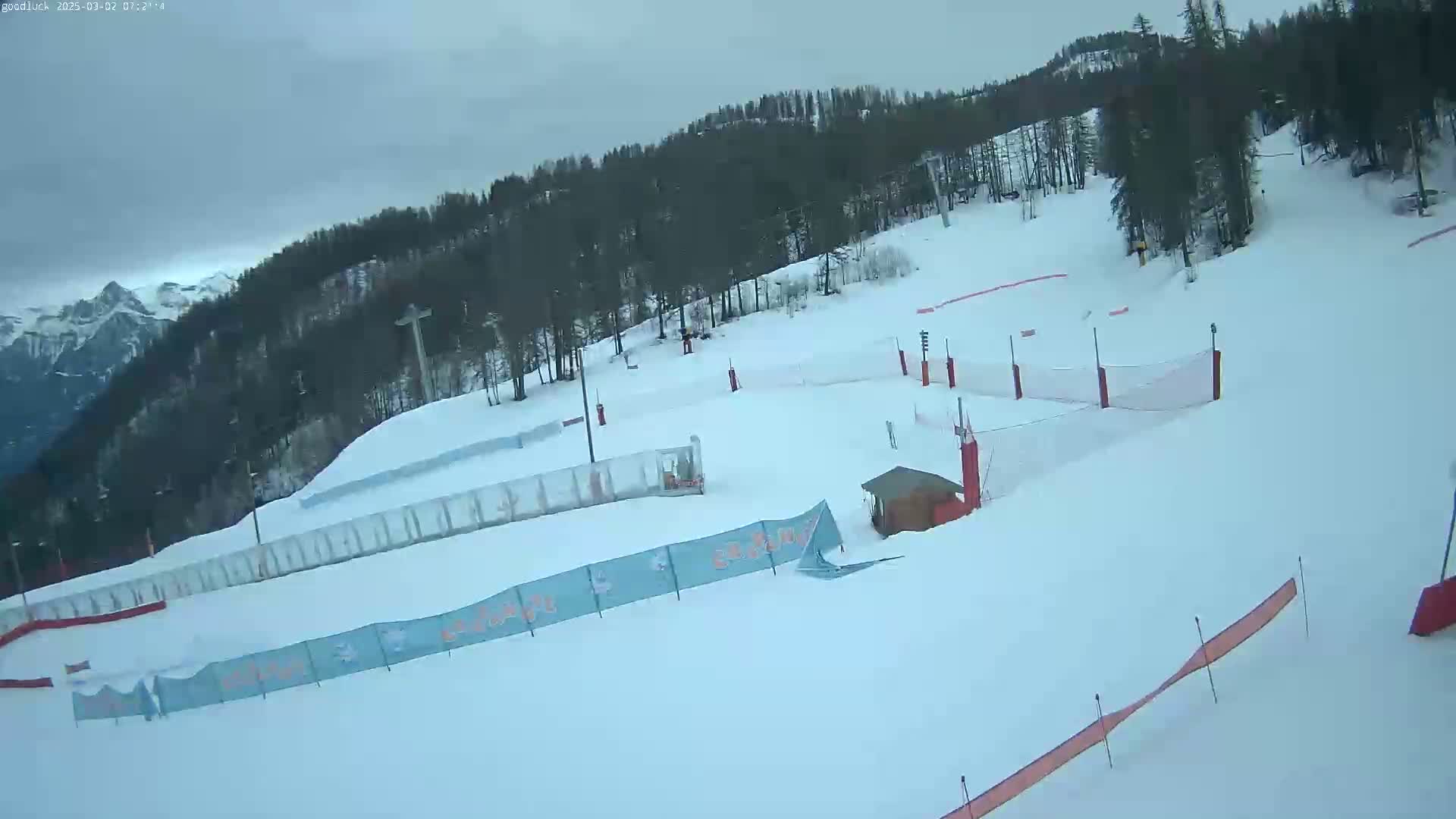 A snow-covered ski slope with several fences and a small building is visible under an overcast sky.