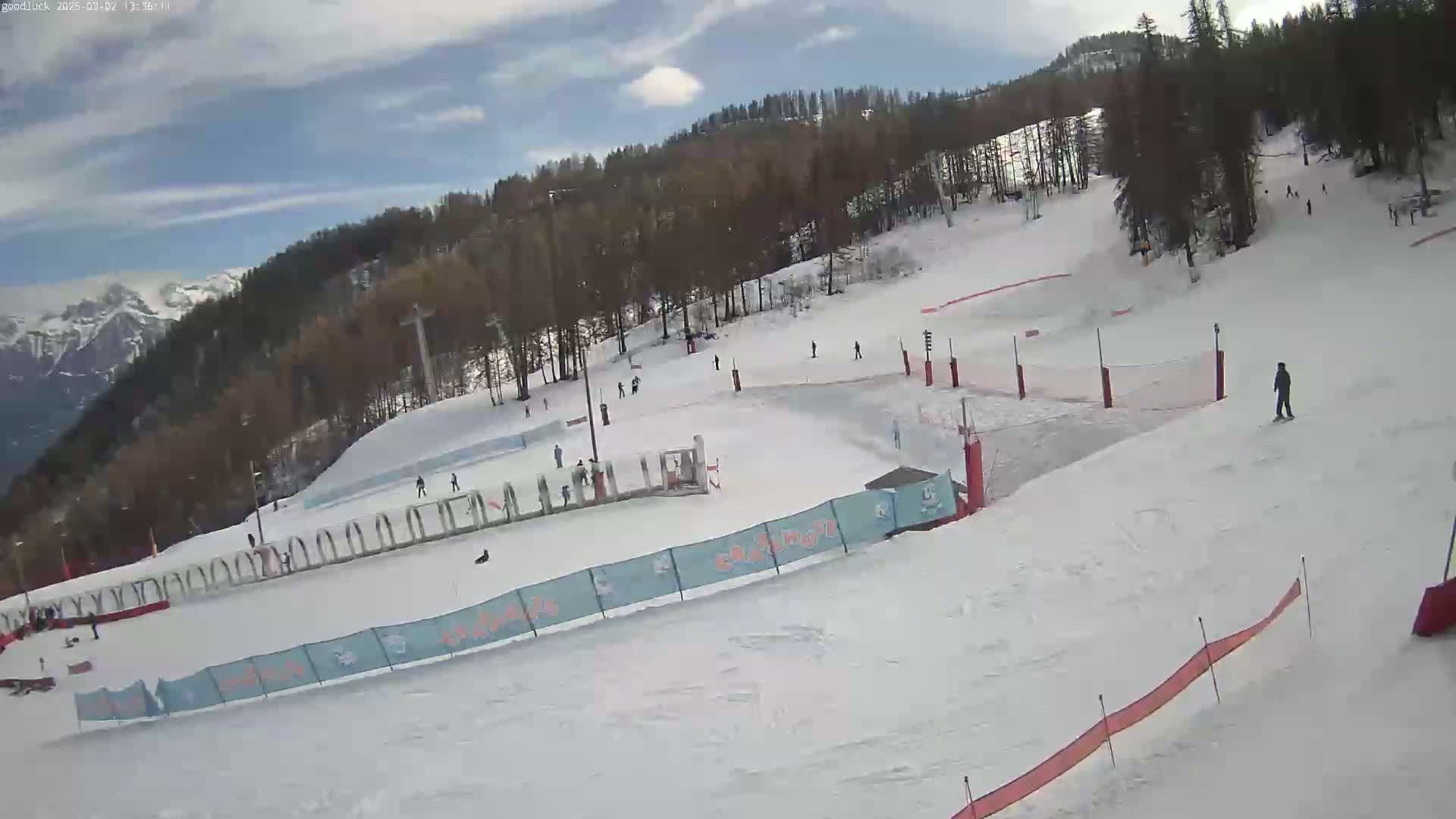 A snow-covered ski slope with several skiers and snowboarders, surrounded by trees and mountains under a partly cloudy sky.