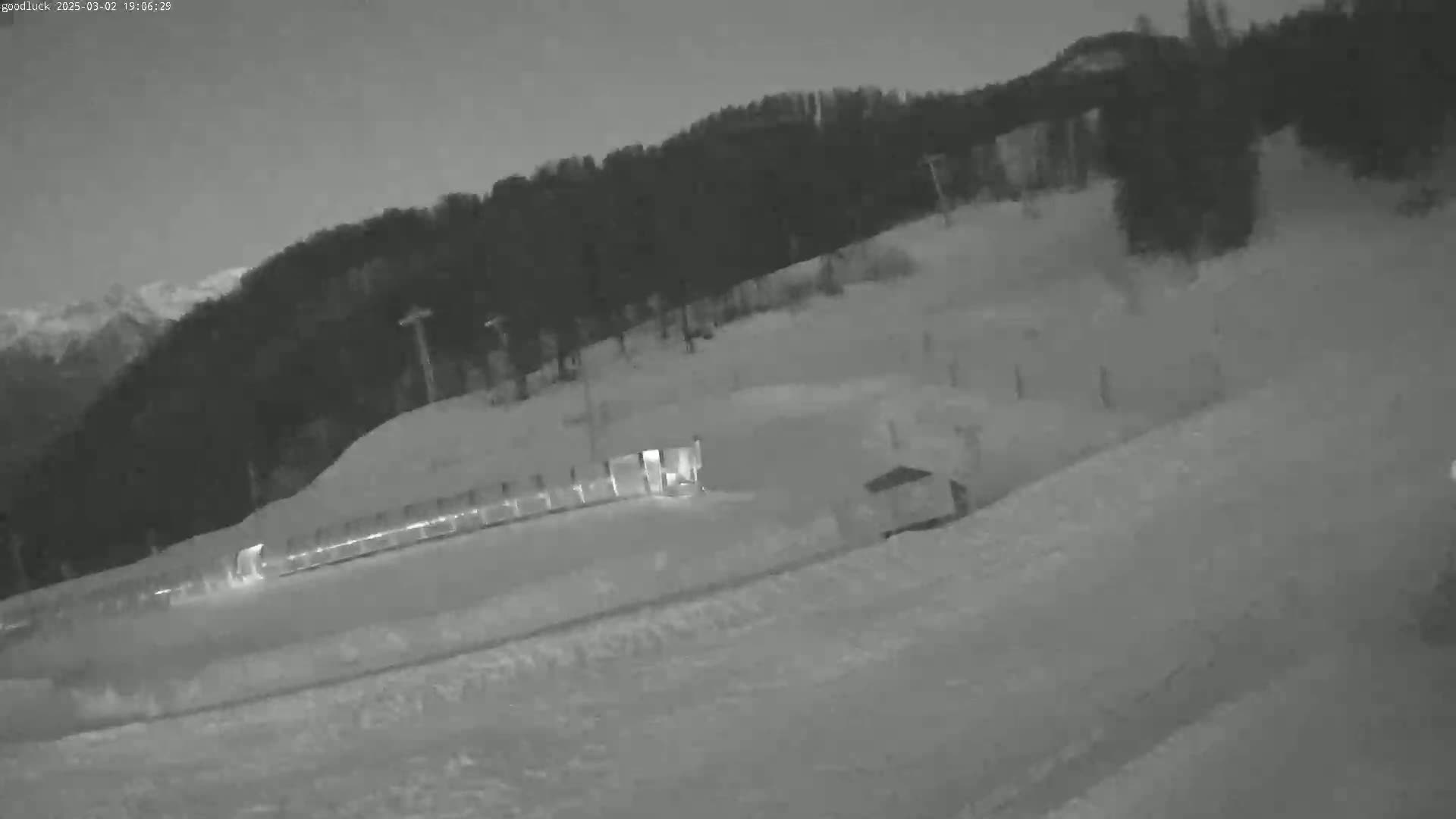 A snow-covered ski slope at night, illuminated by artificial light, with a dark forest in the background under a dark sky.