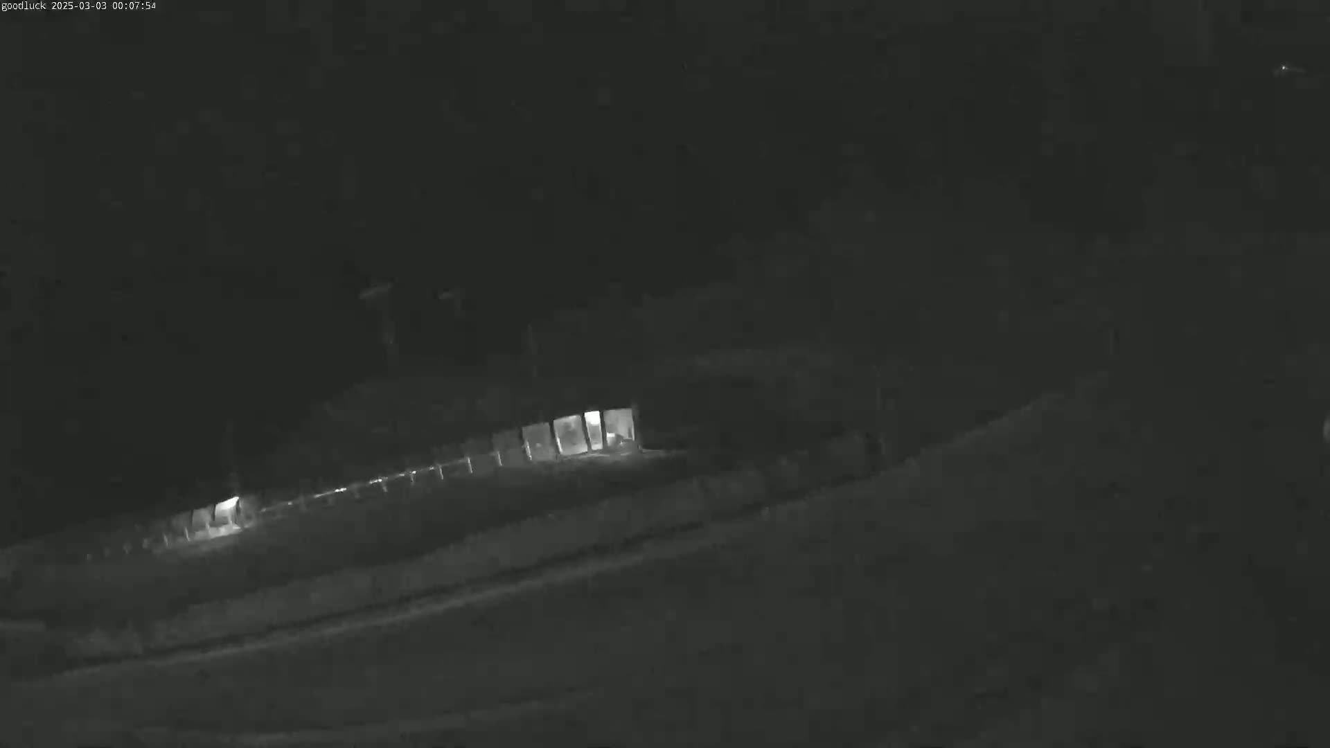 A low-light nighttime image shows a building with illuminated windows and a fence, situated alongside a road.