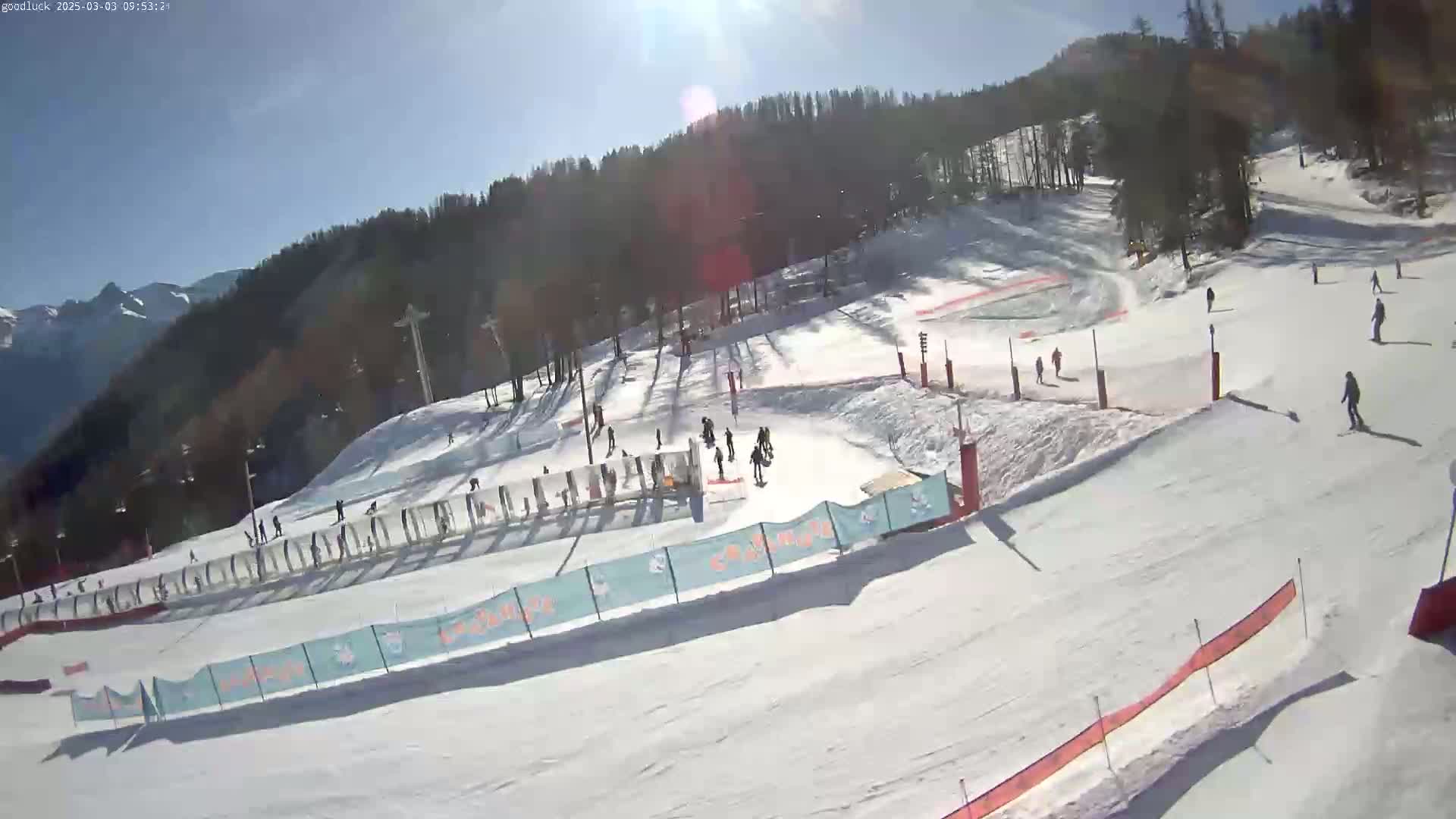 A sunny day at a snow-covered ski slope shows several skiers and snowboarders on the runs, with a mountain range in the background.